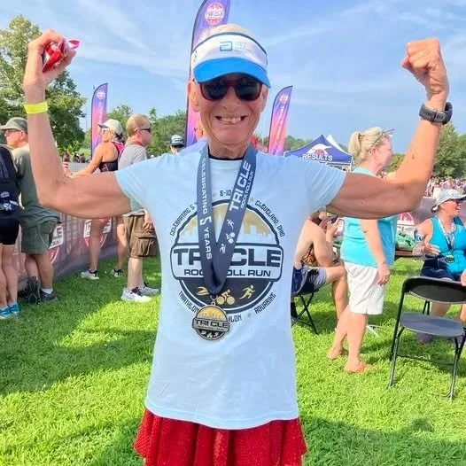 Older woman wearing sunglasses, a blue cap, and a gray T-shirt with a race logo, flexing her arms with a medal around his neck at an outdoor race event with other participants and event tents in the background.