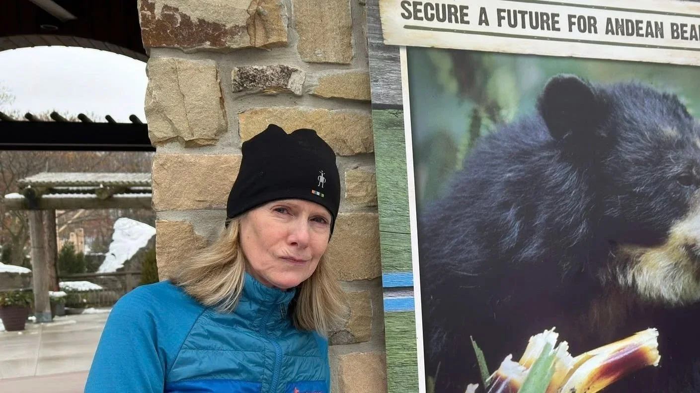 A woman in a blue jacket and black beanie hat stands next to a large bear poster at Andean Bear rescue center. The woman is outdoors with snow on the ground and a stone wall behind her.