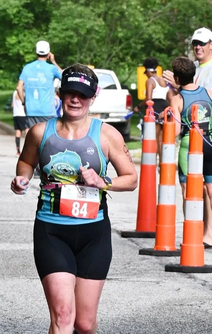 A woman in athletic gear running in a race, wearing a black visor, a gray and blue tank top, black shorts, with the race number 84, and looking determined.