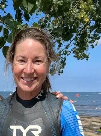 Smiling woman outdoors near the water, wearing sports gear with trees and the ocean in the background.