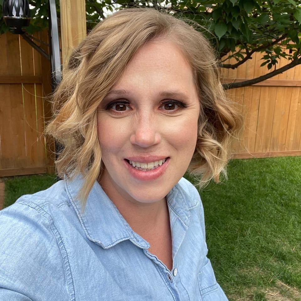A woman with shoulder-length curly blonde hair smiling outdoors in a backyard with a wooden fence, green grass, and a small tree.