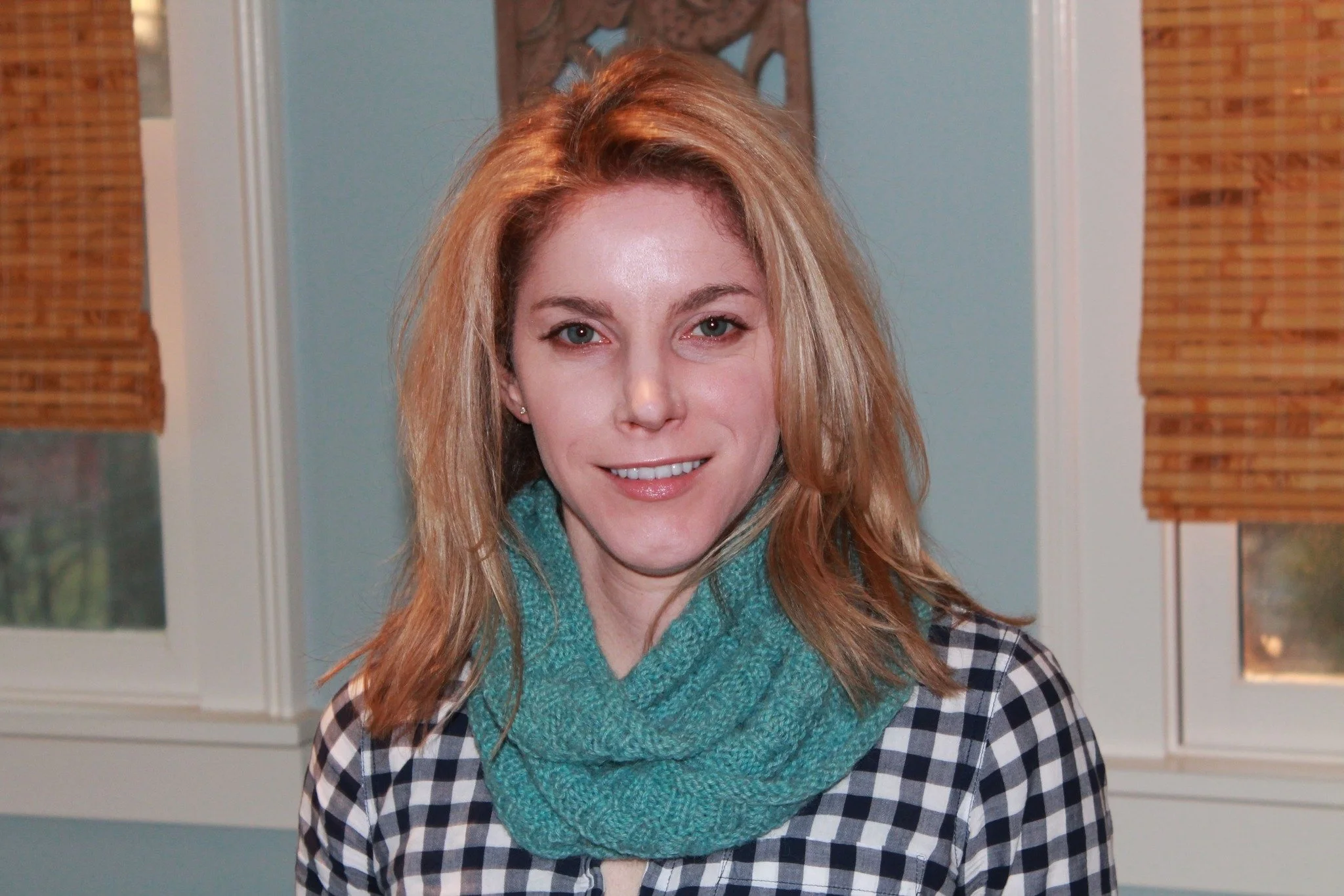 A woman with blonde hair smiling indoors with windows and blinds in the background.
