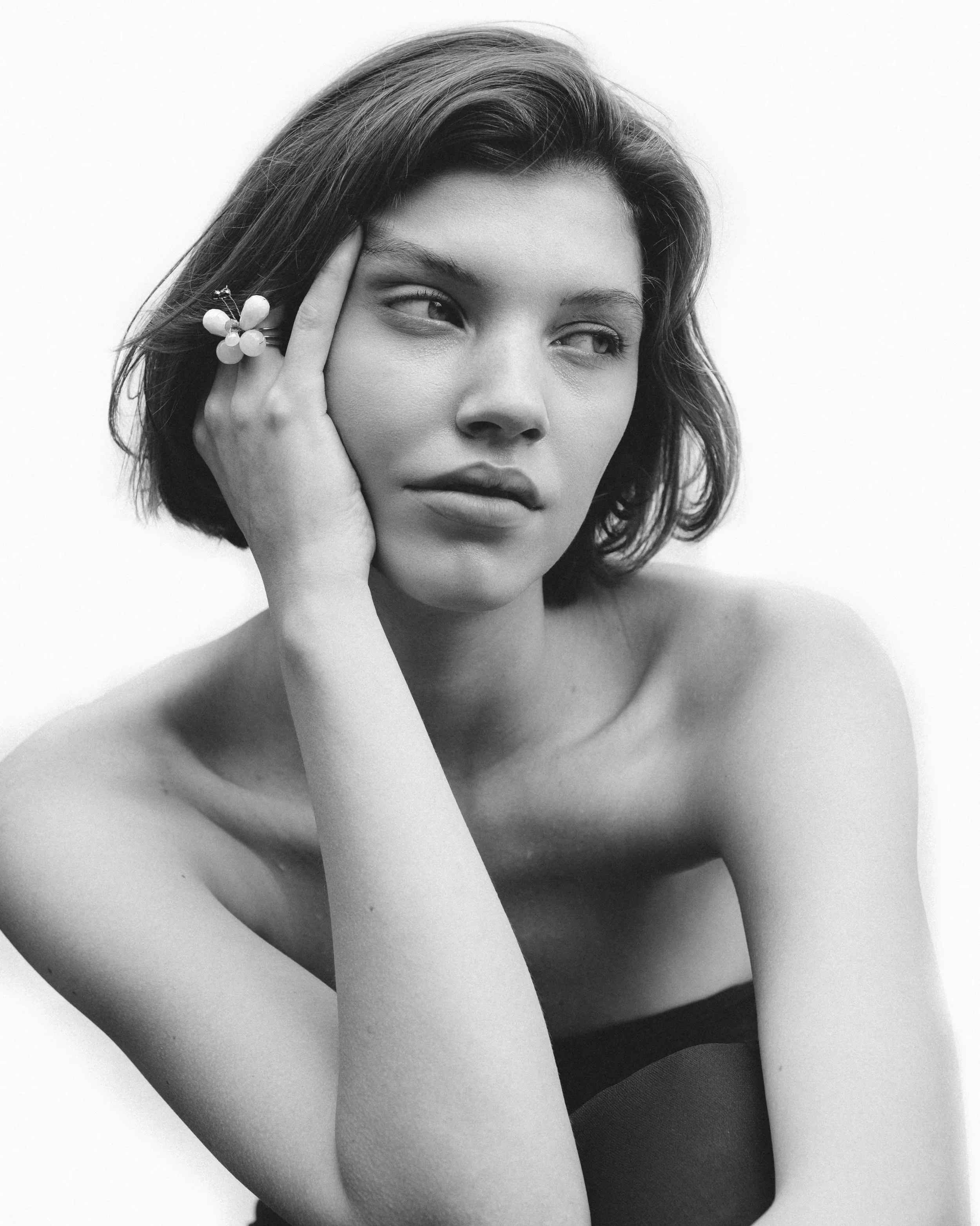 Retrato em preto e branco de uma mulher com cabelo curto, ombros à mostra, fazendo pose pensativa, com a mão apoiada na cabeça ao lado do rosto, uma flor no dedo.