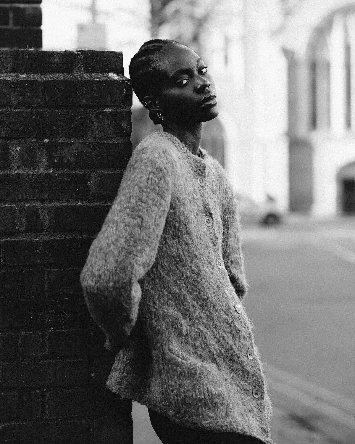 Uma mulher negra com penteado de pequenas tranças, usando uma blusa de tricô com botões, encostada em uma parede de tijolos na rua, em uma foto em preto e branco.