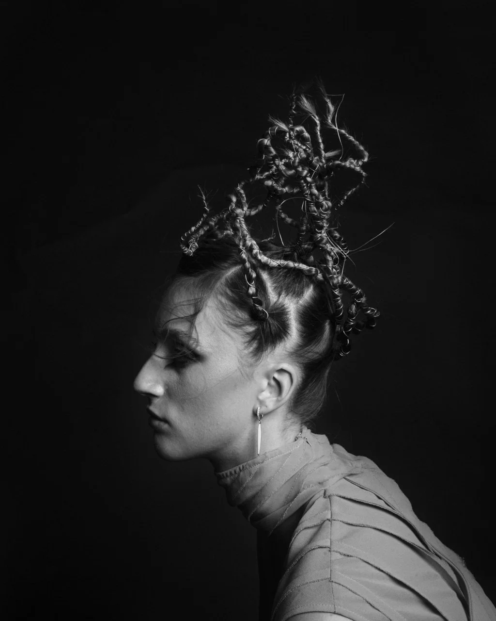 Retrato em preto e branco de uma mulher com cabelo trançado com rolinhos e penteado elaborado, vestindo uma blusa de gola alta e brincos pequenos, de perfil, contra fundo escuro.