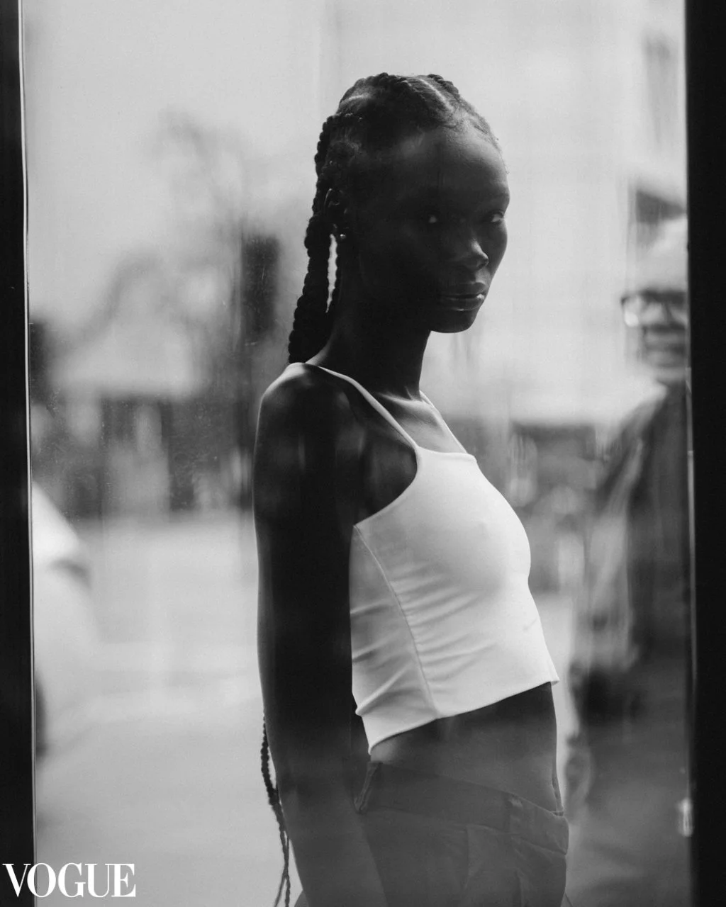 Retrato em preto e branco de uma mulher com penteado de tranças longas, vestindo um top branco e calças escuras, olhando expressivamente para a câmera, com uma parede de vidro ao fundo.