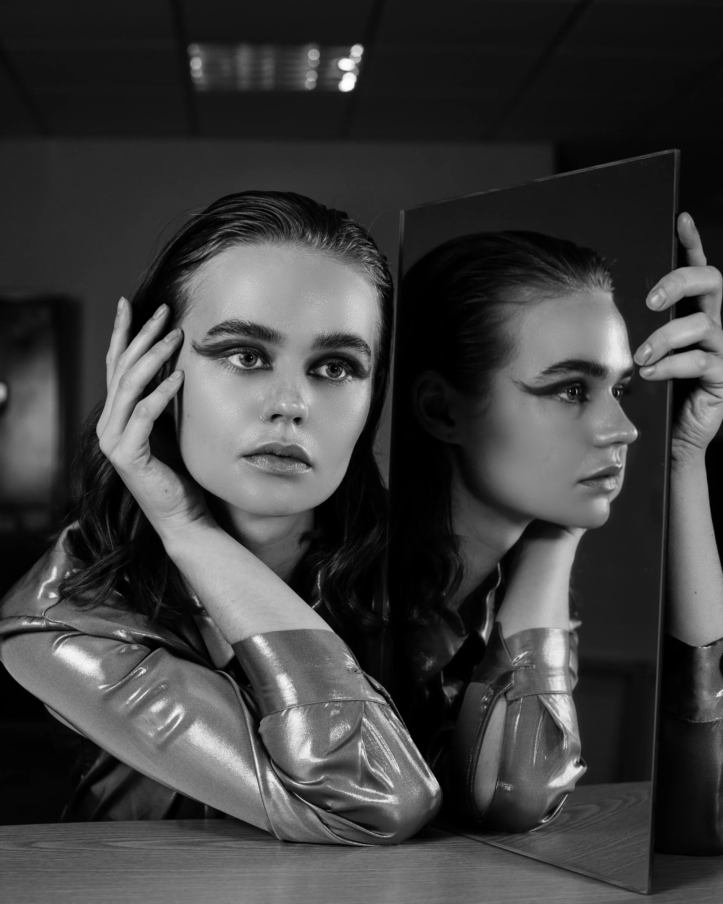 Retrato de uma mulher com cabelo escuro e sobrancelhas grossas, usando uma jaqueta de couro, olhando para uma reflexão no espelho, em uma foto em preto e branco.