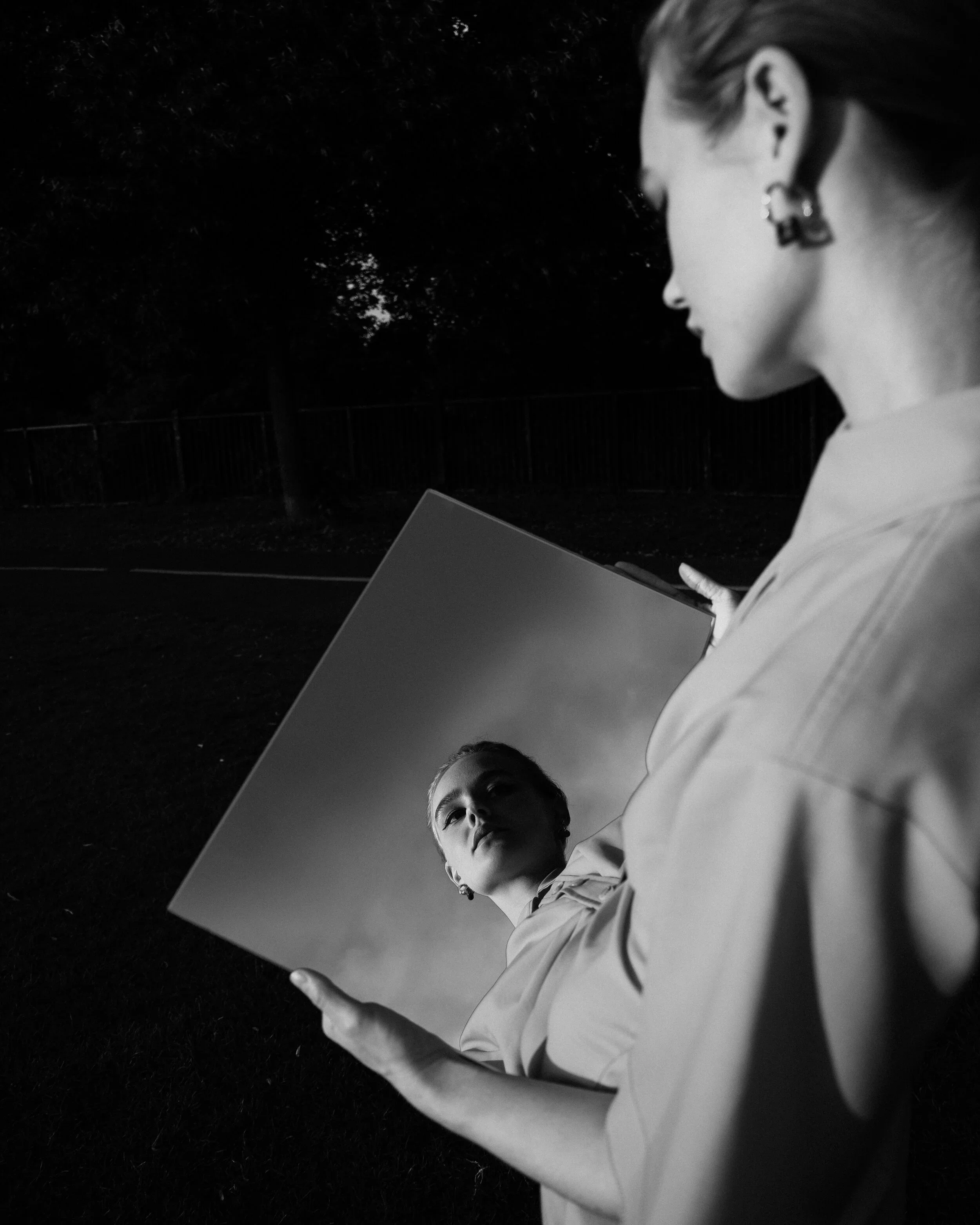 Pessoa olhando para um espelho ao ar livre à noite, refletindo seu rosto. Em preto e branco.