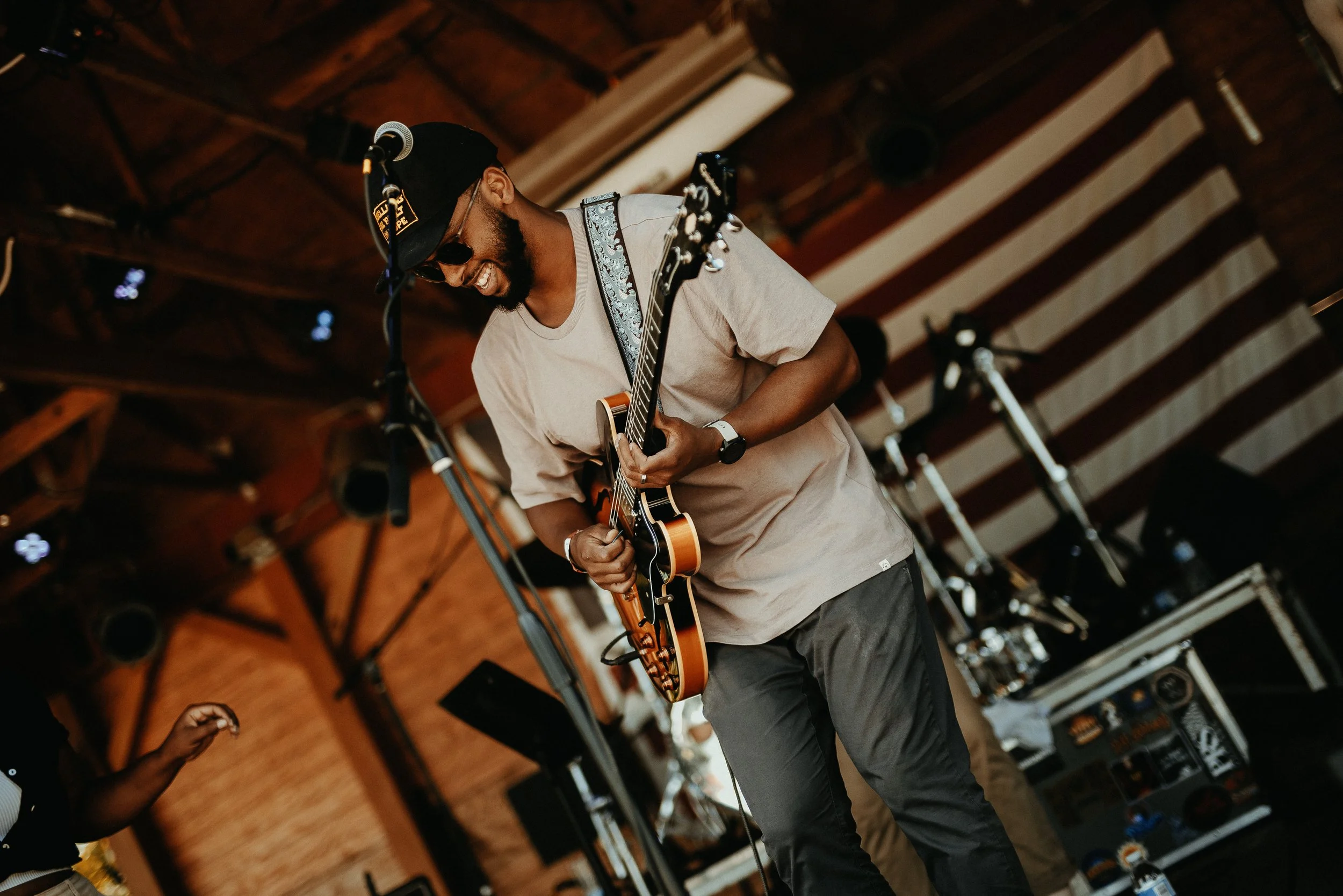 Man playing an electric guitar on stage, wearing sunglasses, a beige t-shirt, a black cap, and a wristwatch, smiling, with a microphone and stage equipment visible in the background.
