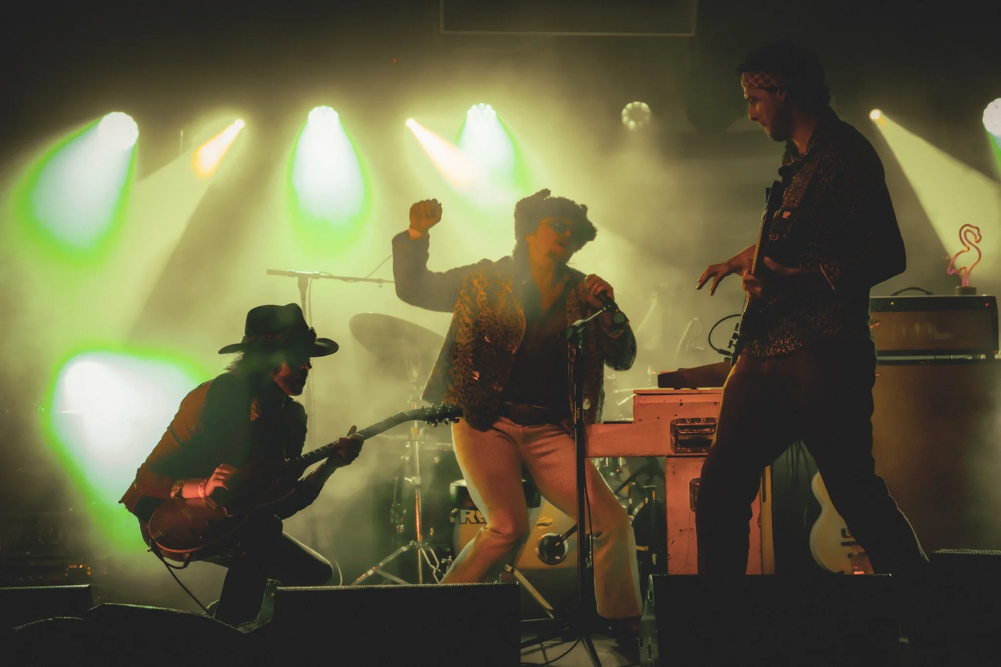 Musicians performing on stage with colorful lights, including a person singing into a microphone, a guitarist, and a keyboard player, surrounded by stage equipment.