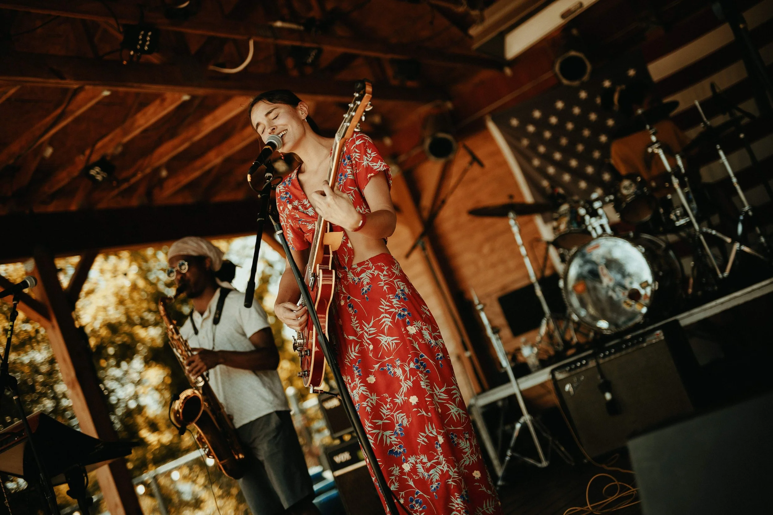 A woman in a red floral dress playing an electric guitar and singing into a microphone on a stage with a wooden roof, accompanied by a man in a white shirt and headscarf playing a saxophone and a drum set in the background.