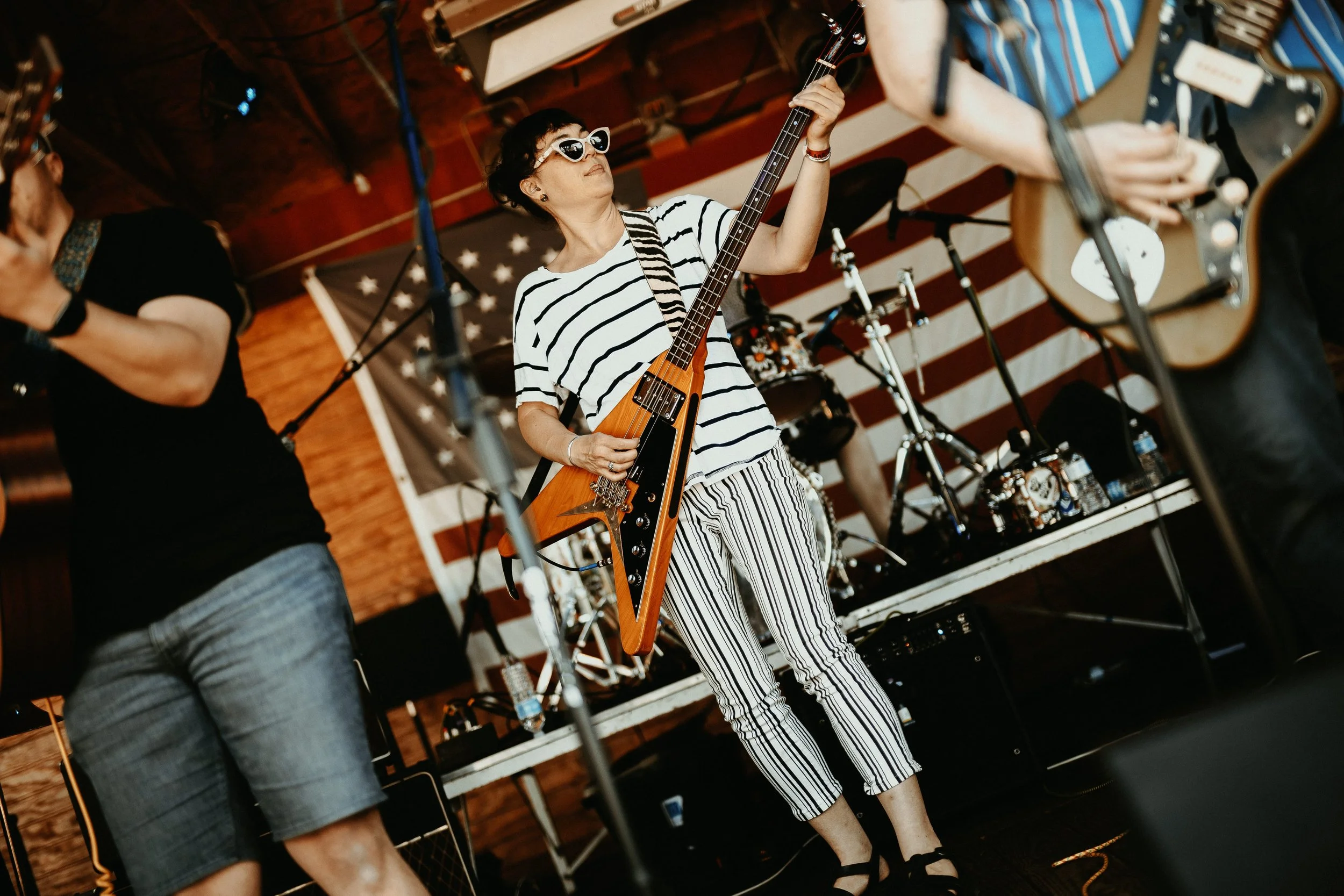 A woman wearing sunglasses, a striped shirt, and striped pants playing an electric guitar on stage during a live music performance with a band.