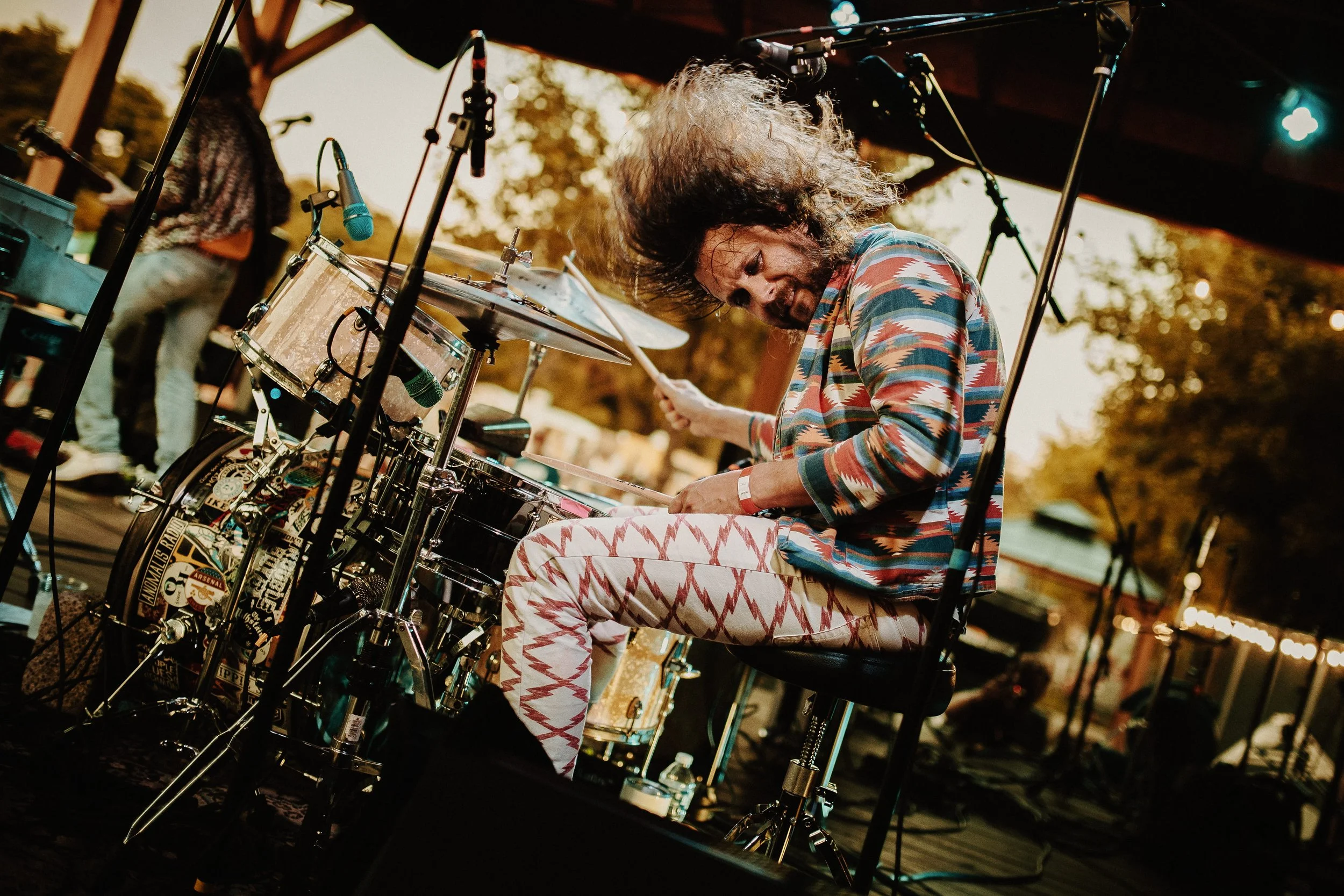 A man with long curly hair, wearing a colorful patterned shirt and white pants with red zigzag patterns, is playing a drum set on an outdoor stage during sunset. His hair is flying as he strikes the drums energetically.