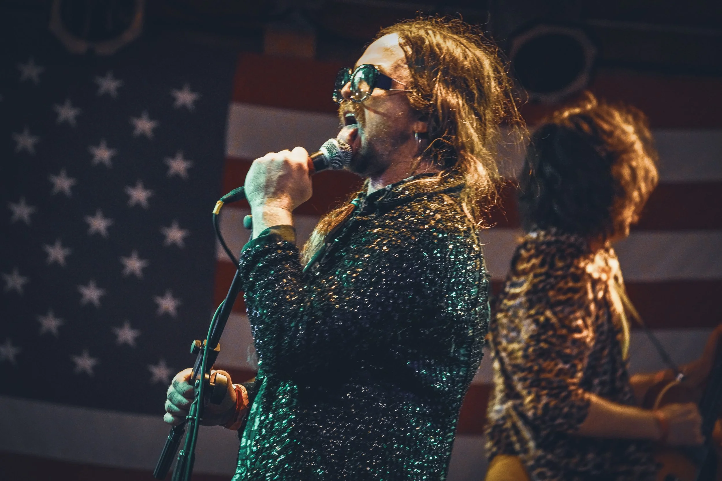 A singer with dreadlocks, sunglasses, and a sequined jacket singing into a microphone, with an American flag in the background.