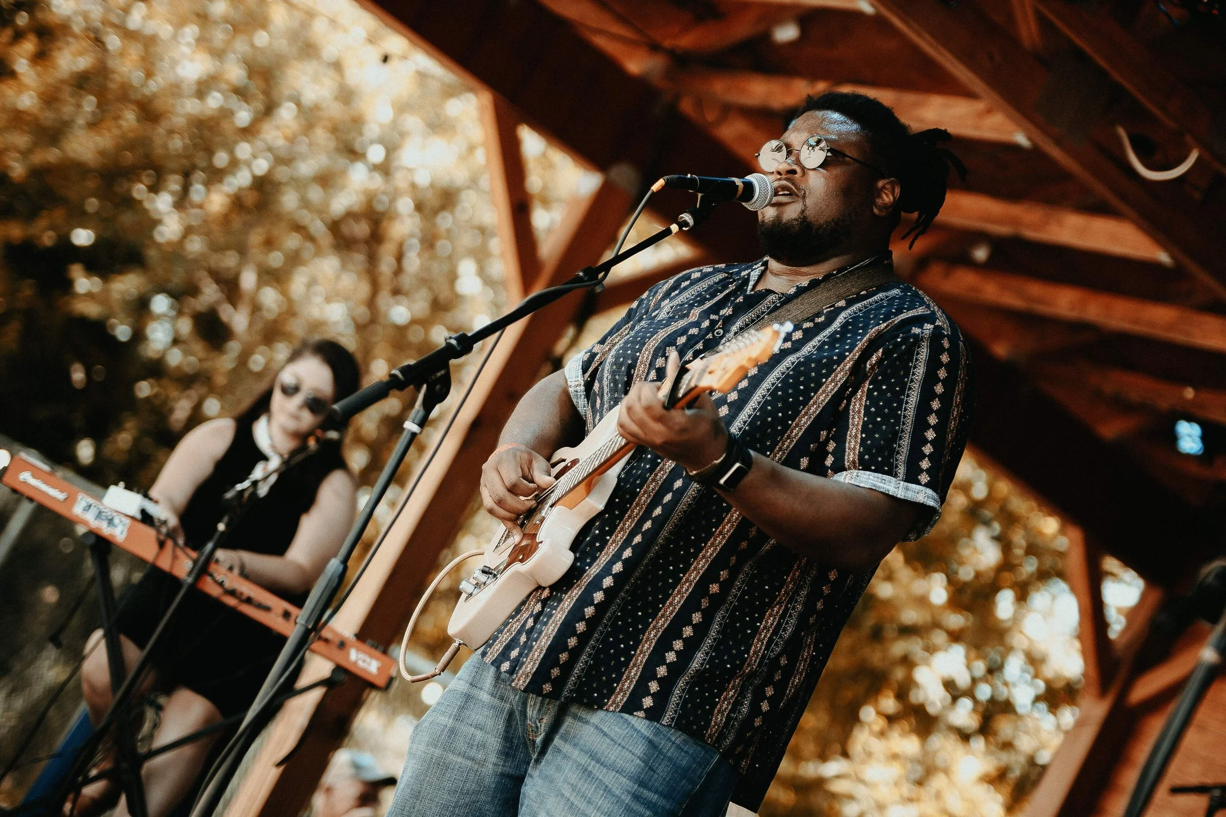 A man with dreadlocks and glasses playing an electric guitar and singing into a microphone during an outdoor performance, with a woman in the background using a keyboard.
