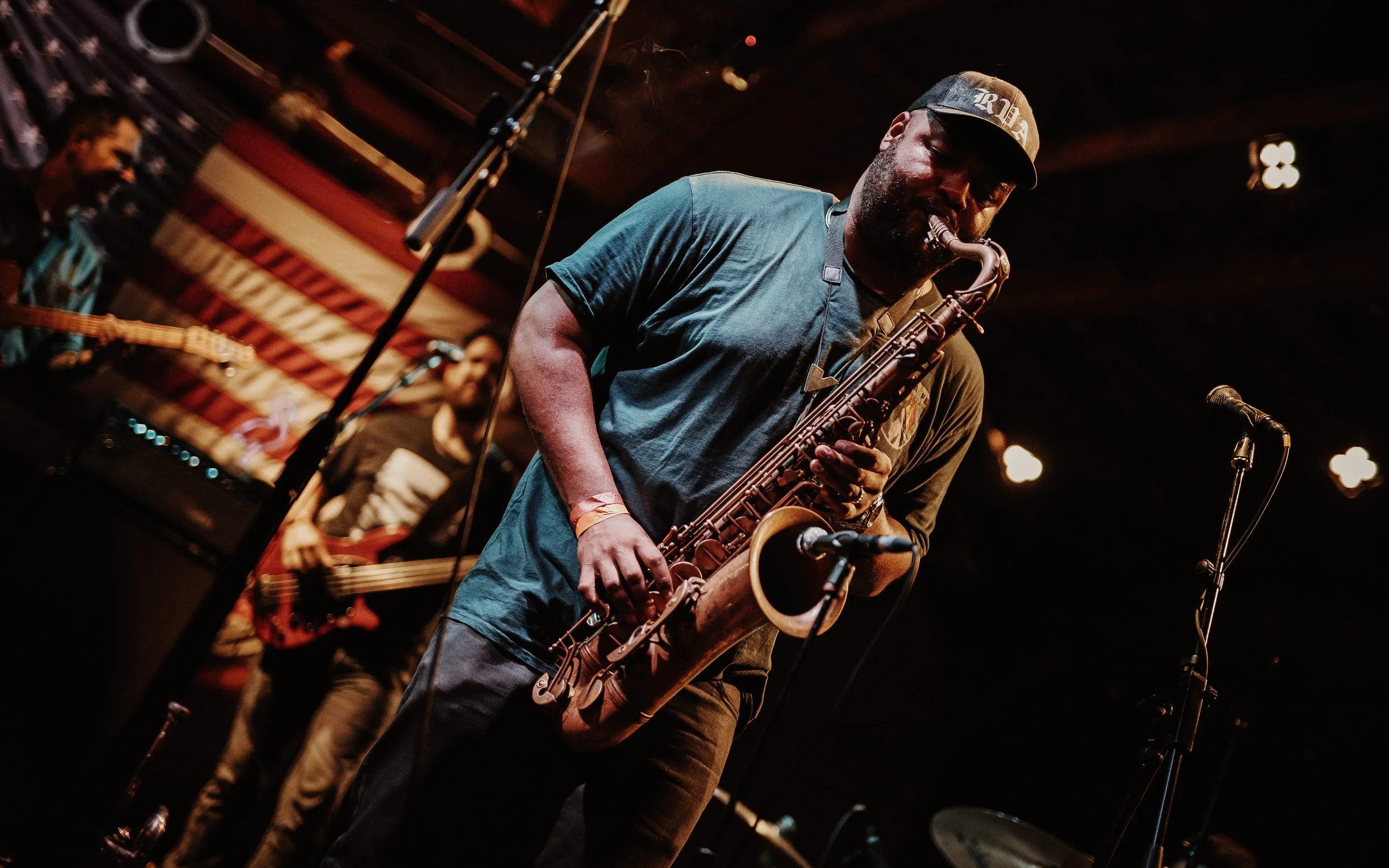 A man playing the saxophone on stage with a band in the background, at a live music venue.
