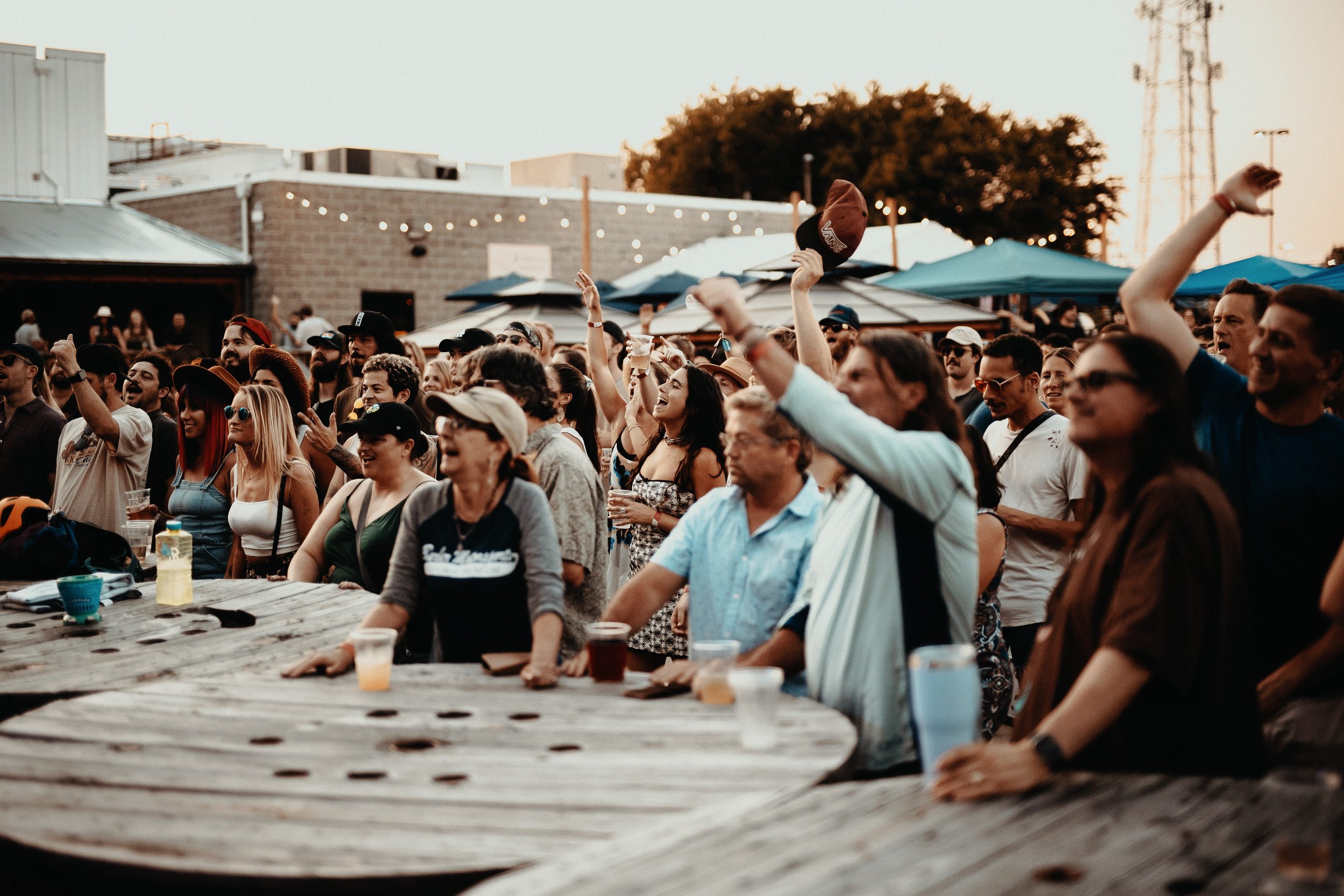 A crowd of people enjoying an outdoor concert or event around sunset, with some raising their hands and holding drinks, featuring a wooden table in the foreground and tents in the background.