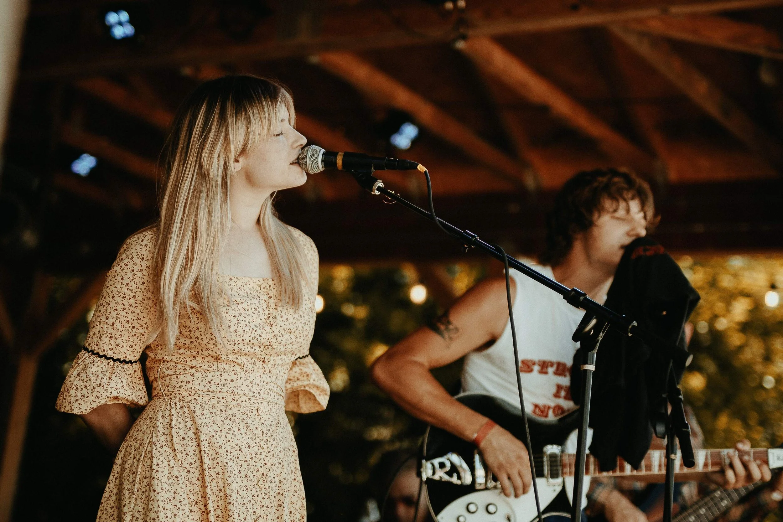 A young woman with blonde hair singing into a microphone and a young man with curly hair playing an electric guitar during a live performance under a wooden pavilion.