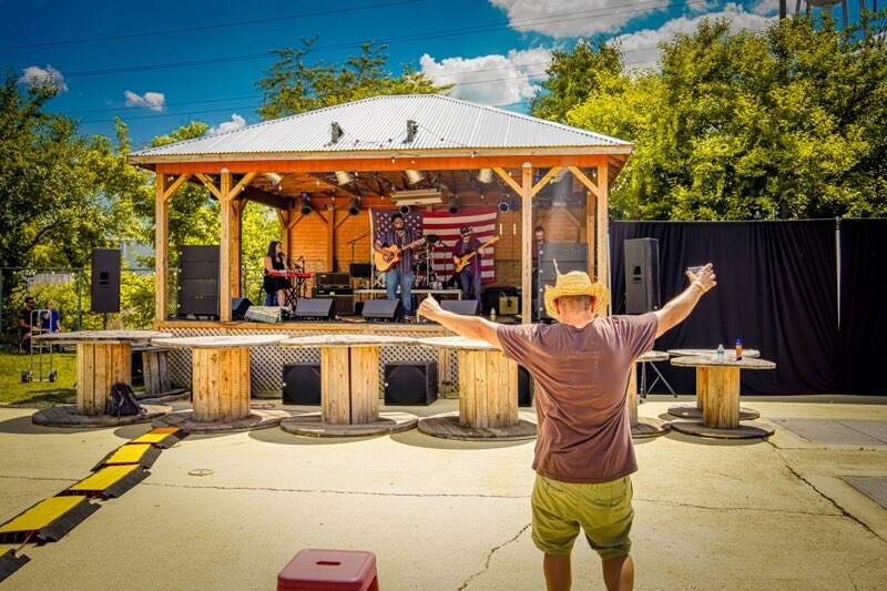 A person wearing a cowboy hat with arms outstretched, facing a wooden stage with a band performing. The band includes a guitarist, a keyboardist, and a bassist, with an American flag hanging in the background. The setting is outdoors on a sunny day w