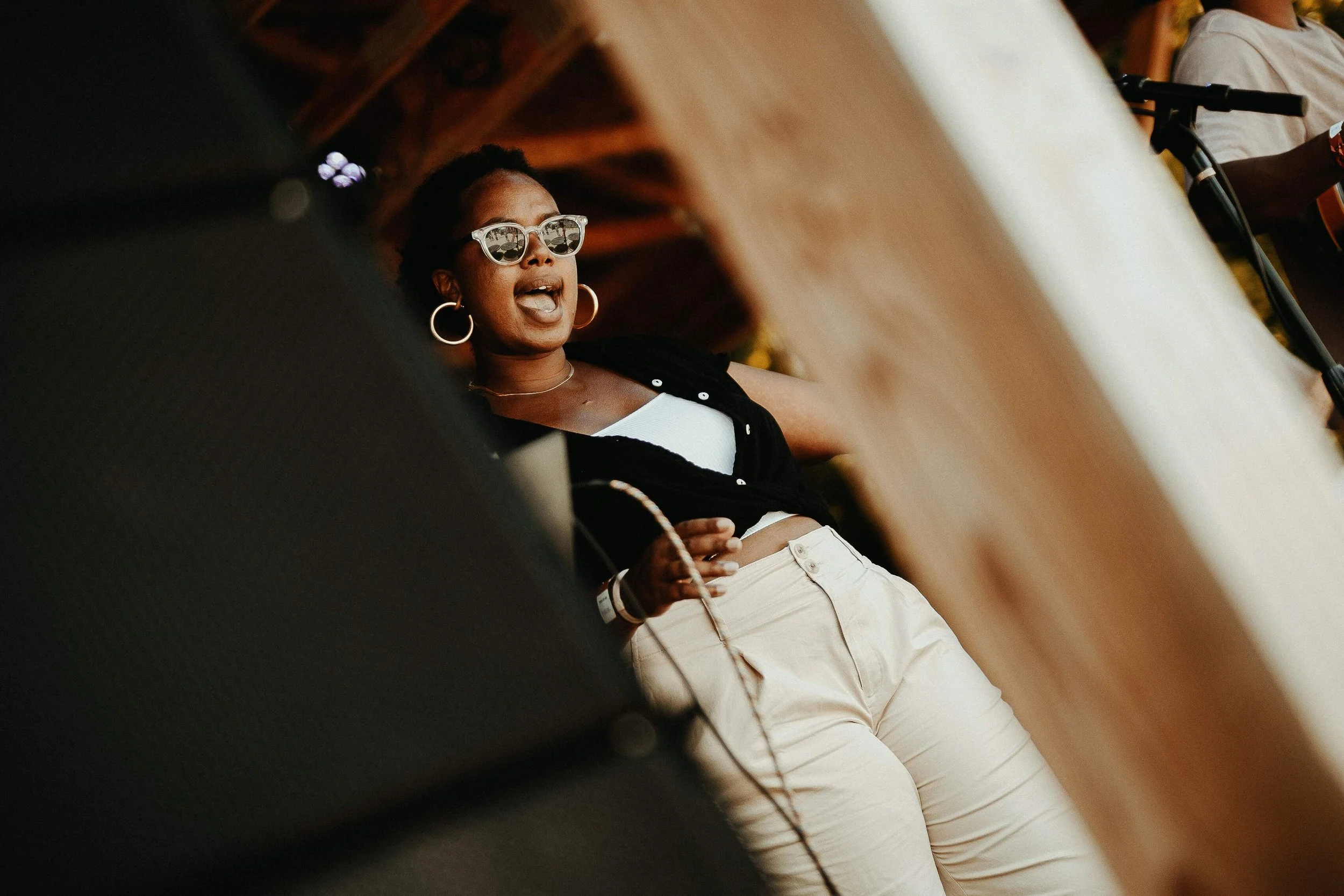 A woman with dark skin wearing sunglasses, hoop earrings, a black top over a white crop top, and beige pants, is talking or singing during an outdoor event, with people and a microphone stand in the background.