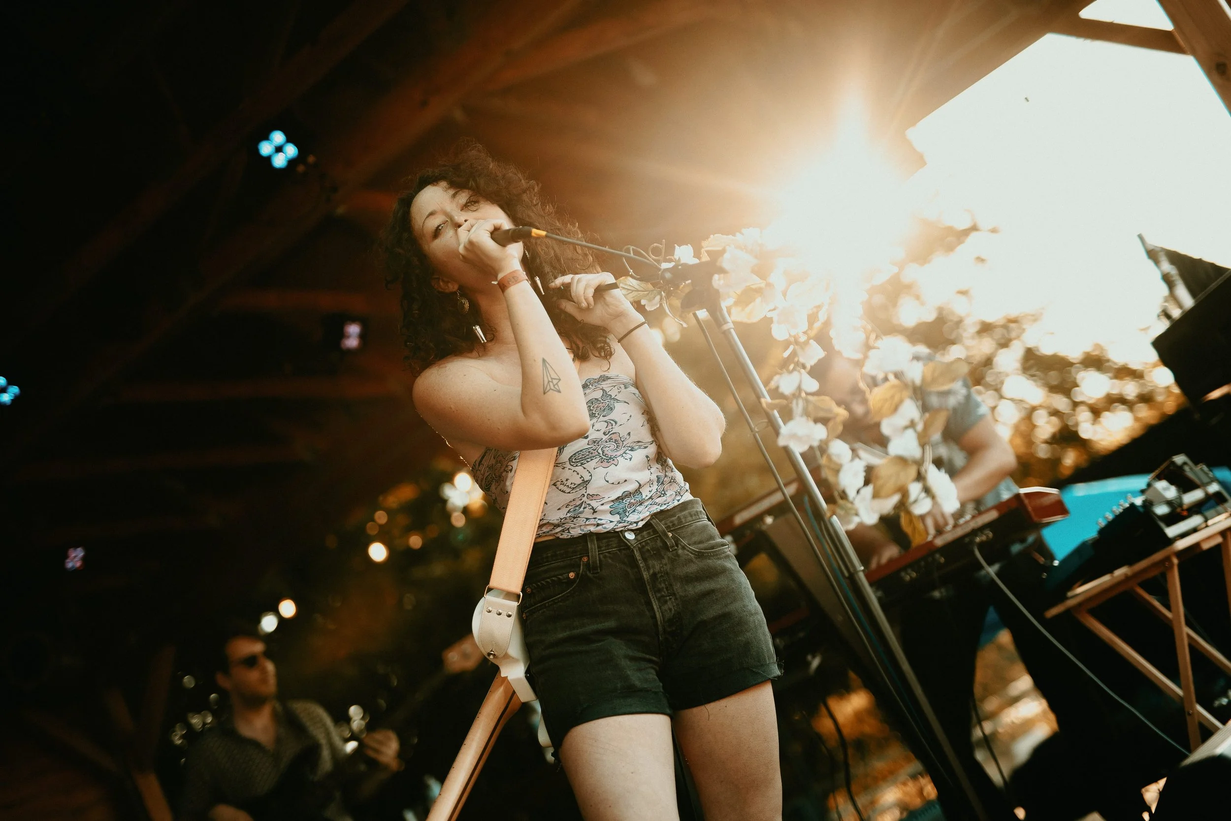 A woman singing into a microphone at an outdoor music event during sunset, with a keyboard and a man in the background playing guitar.