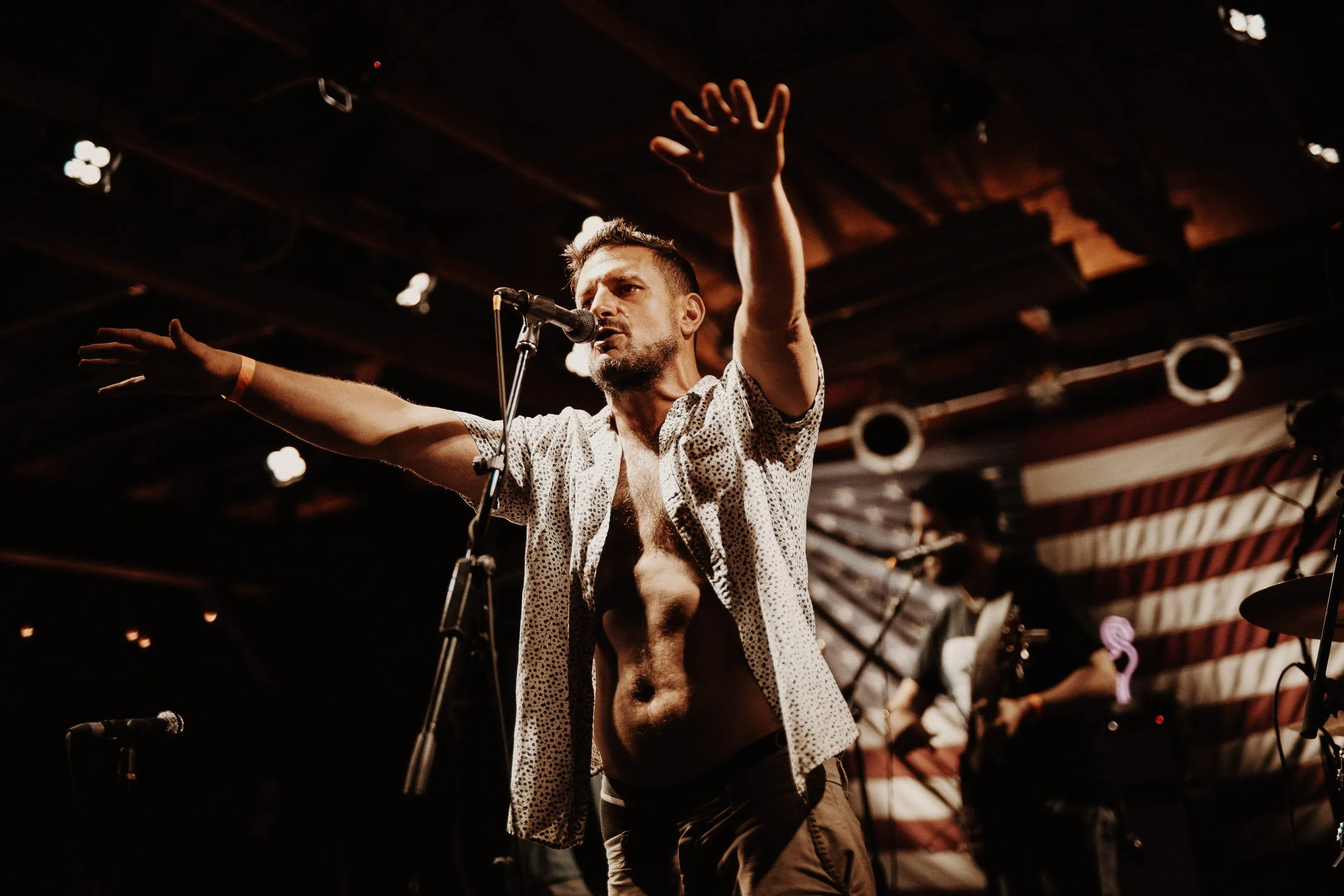 Male performer on stage with arms outstretched, speaking or singing into a microphone. Background includes band members and American flag banner. Dim stage lighting creates a dramatic atmosphere.