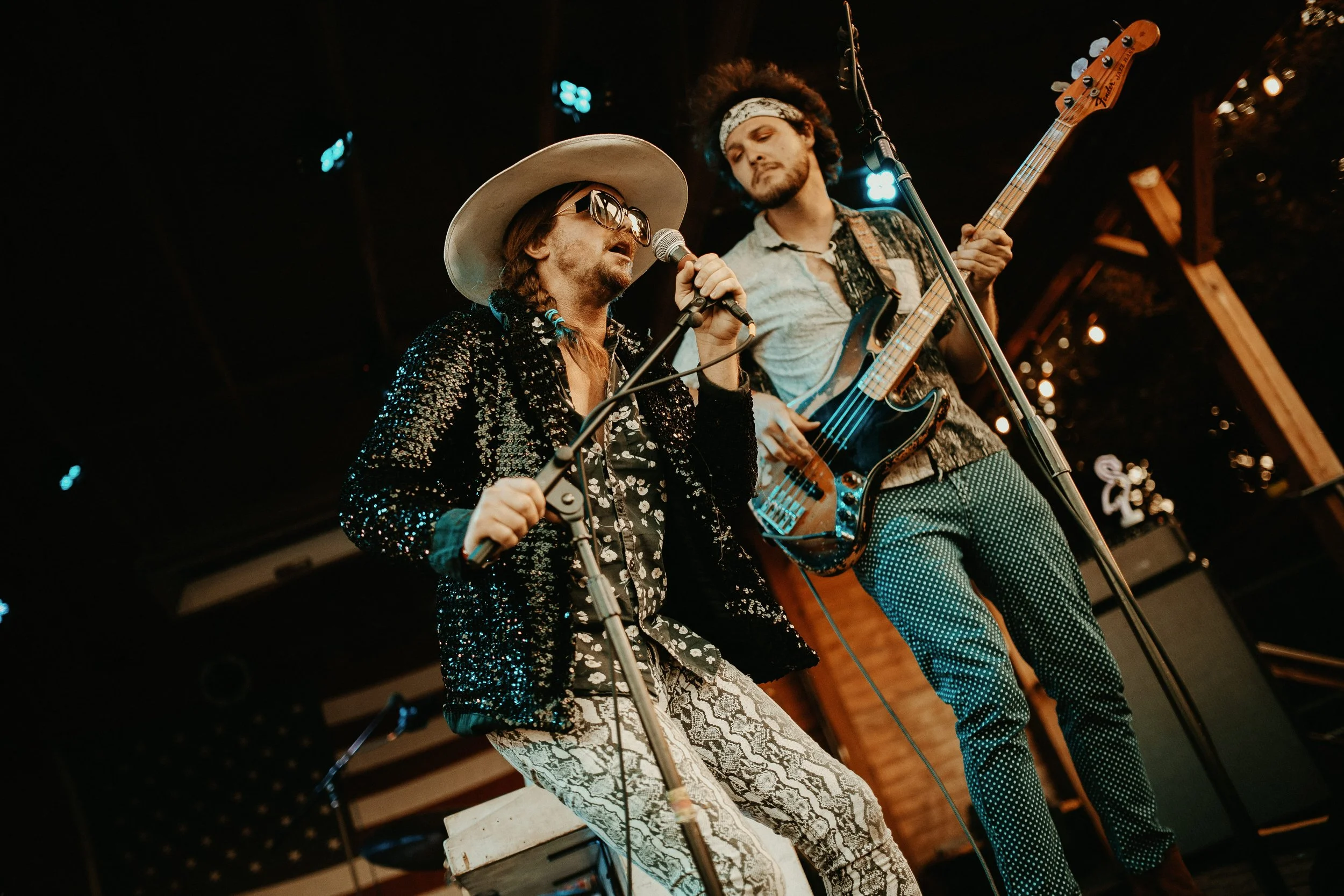 Two male musicians performing on stage, one singing into a microphone and wearing sunglasses, a wide-brimmed hat, and a sparkly jacket, the other playing an electric bass guitar, with curly hair and a patterned bandana, both dressed in vintage-style 