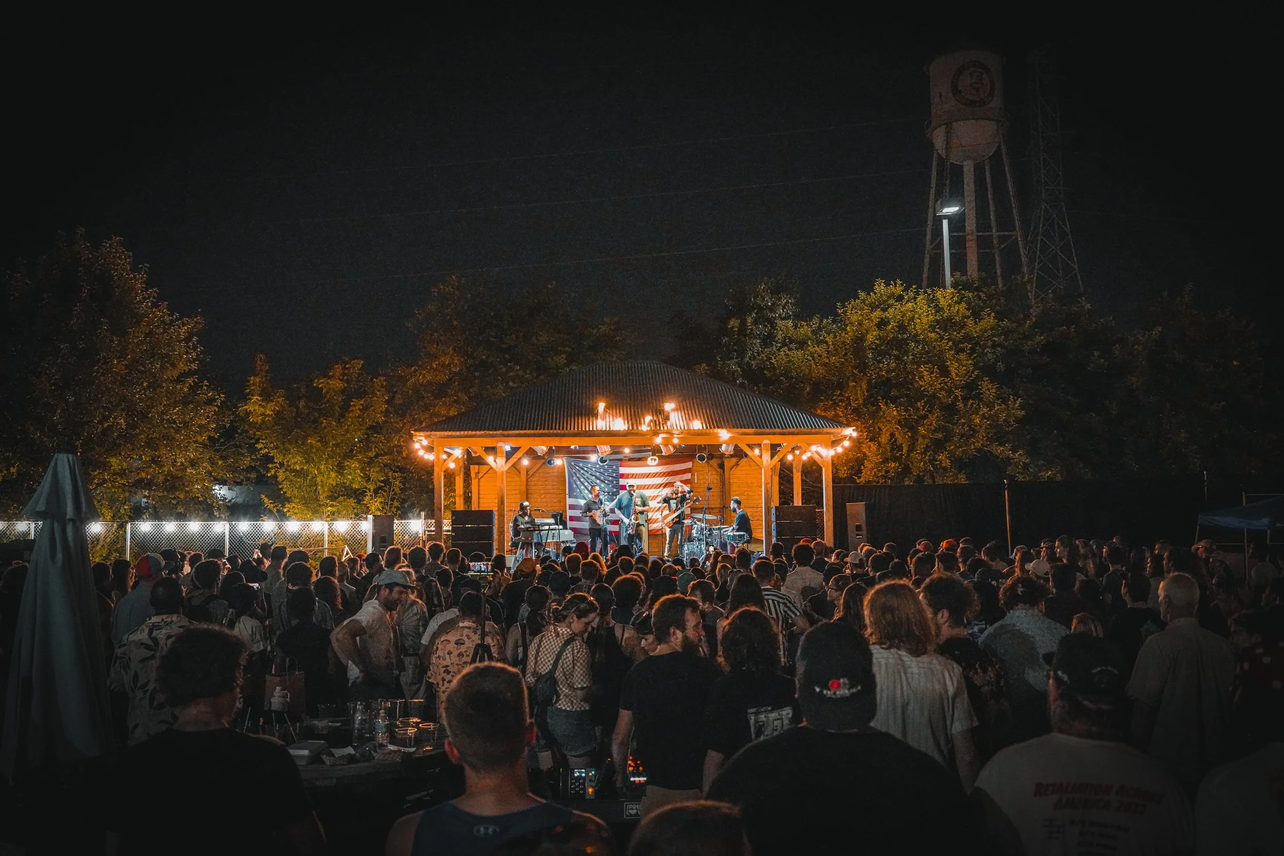 An outdoor concert at night with a band performing on a wooden stage decorated with string lights and an American flag. A large crowd listens, surrounded by trees and industrial water towers.