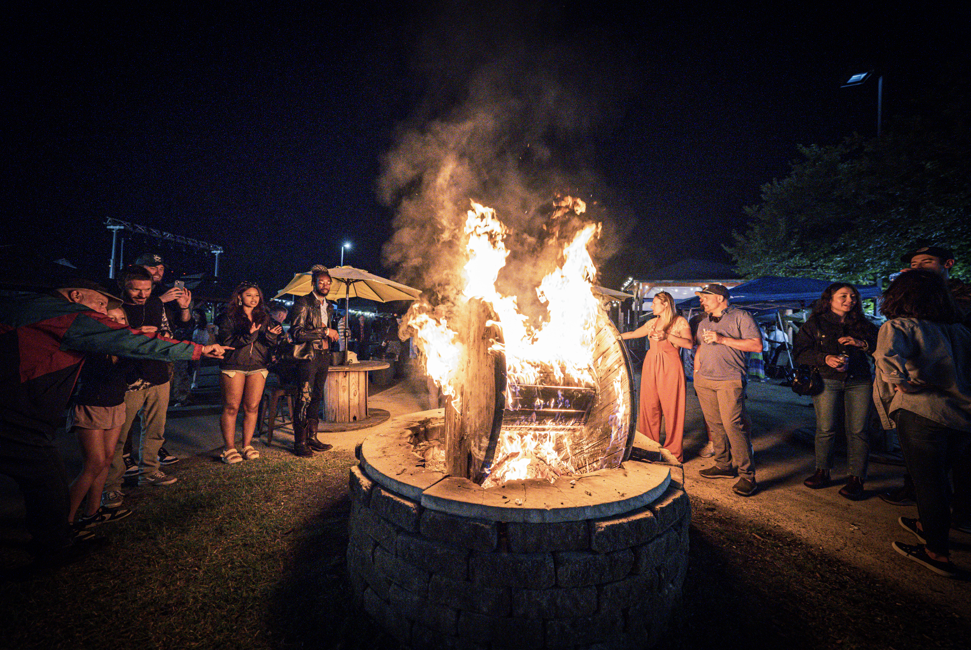 People gathered around a large, open fire pit at night, with some taking pictures or watching the flames, and others talking. The fire is contained within a round stone structure, and flames are rising high, illuminating the nighttime scene.