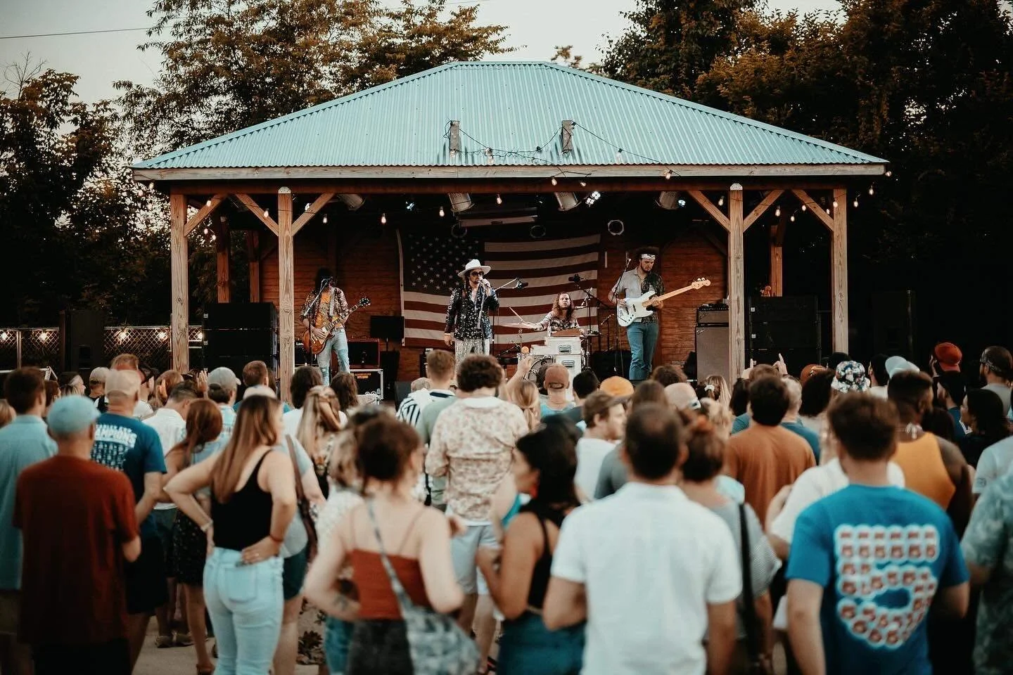 Outdoor concert with a band performing on a wooden stage, large American flag backdrop, and audience watching.