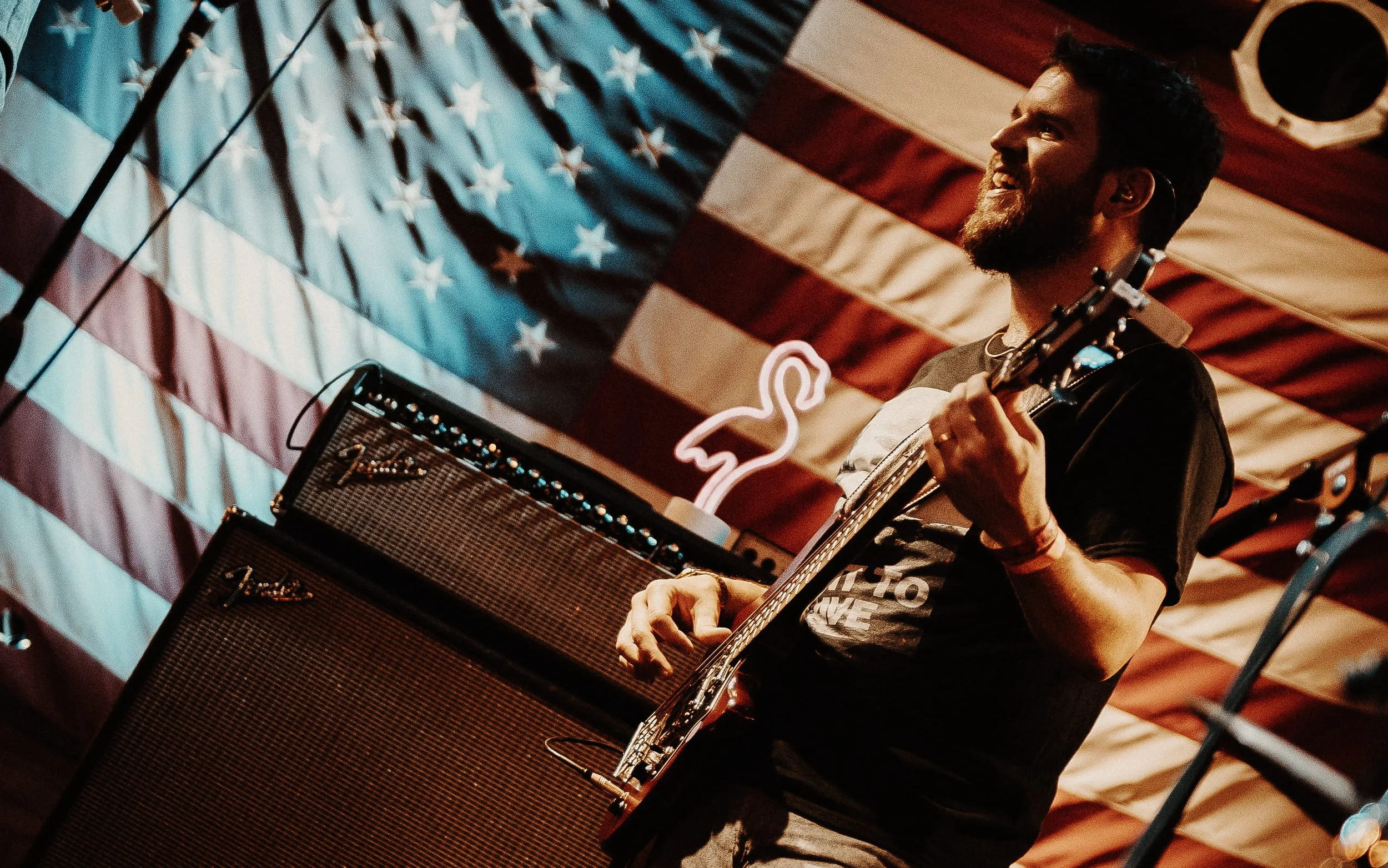 Musician playing electric guitar on stage with an American flag backdrop and neon flamingo sign.