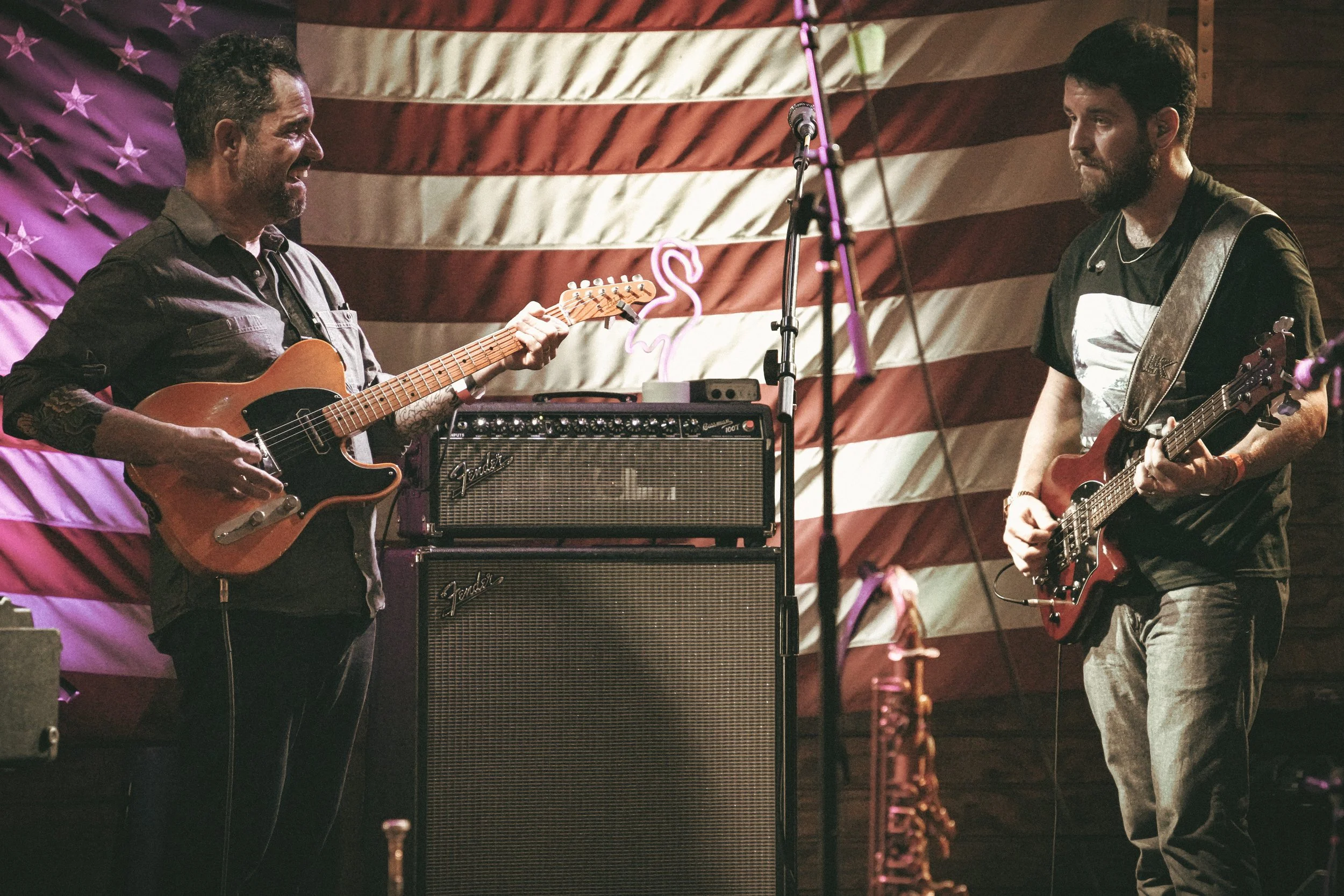 Two musicians playing electric guitars on stage with an American flag backdrop, a neon flamingo light, and musical equipment in the background.