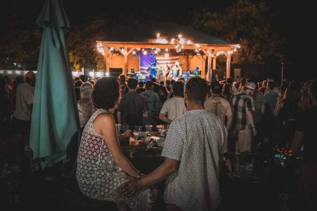 Outdoor concert scene at night with a singing band on stage illuminated with string lights, crowd gathered in front, including a couple holding hands in the foreground.
