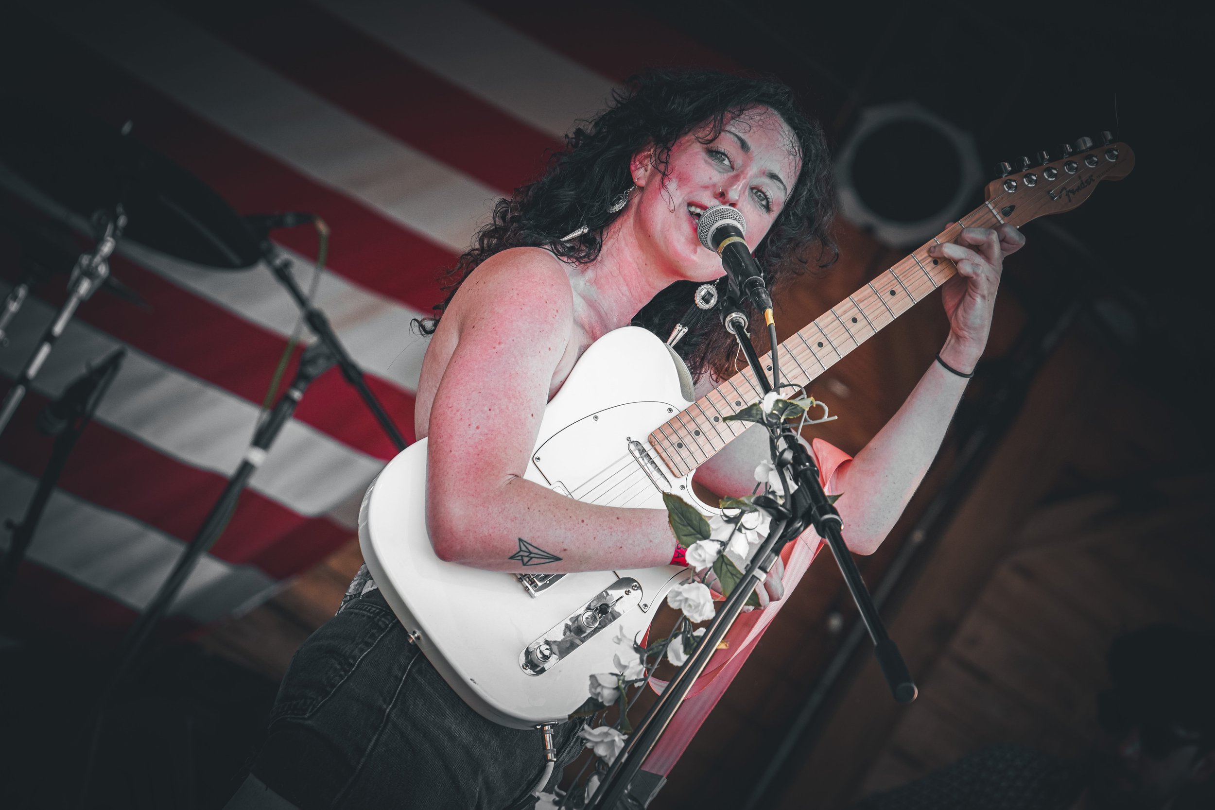 A woman singing and playing an electric guitar on stage with a microphone, decorated with artificial flowers, in front of an American flag backdrop.