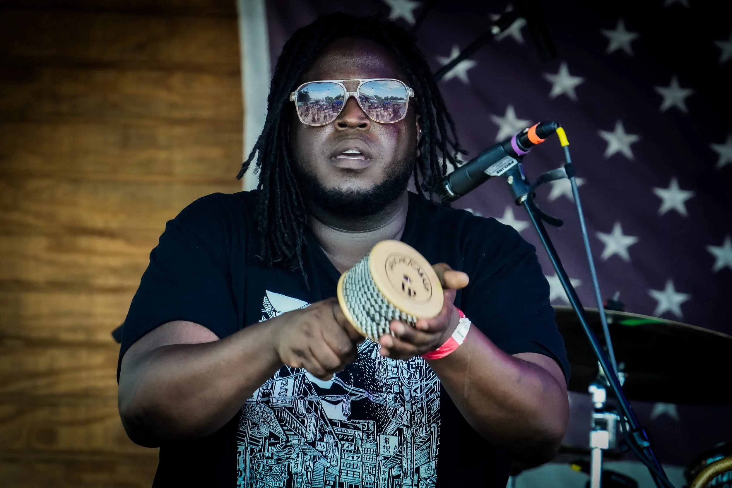 Man with sunglasses and dreadlocks playing a percussion instrument on stage with American flag backdrop.