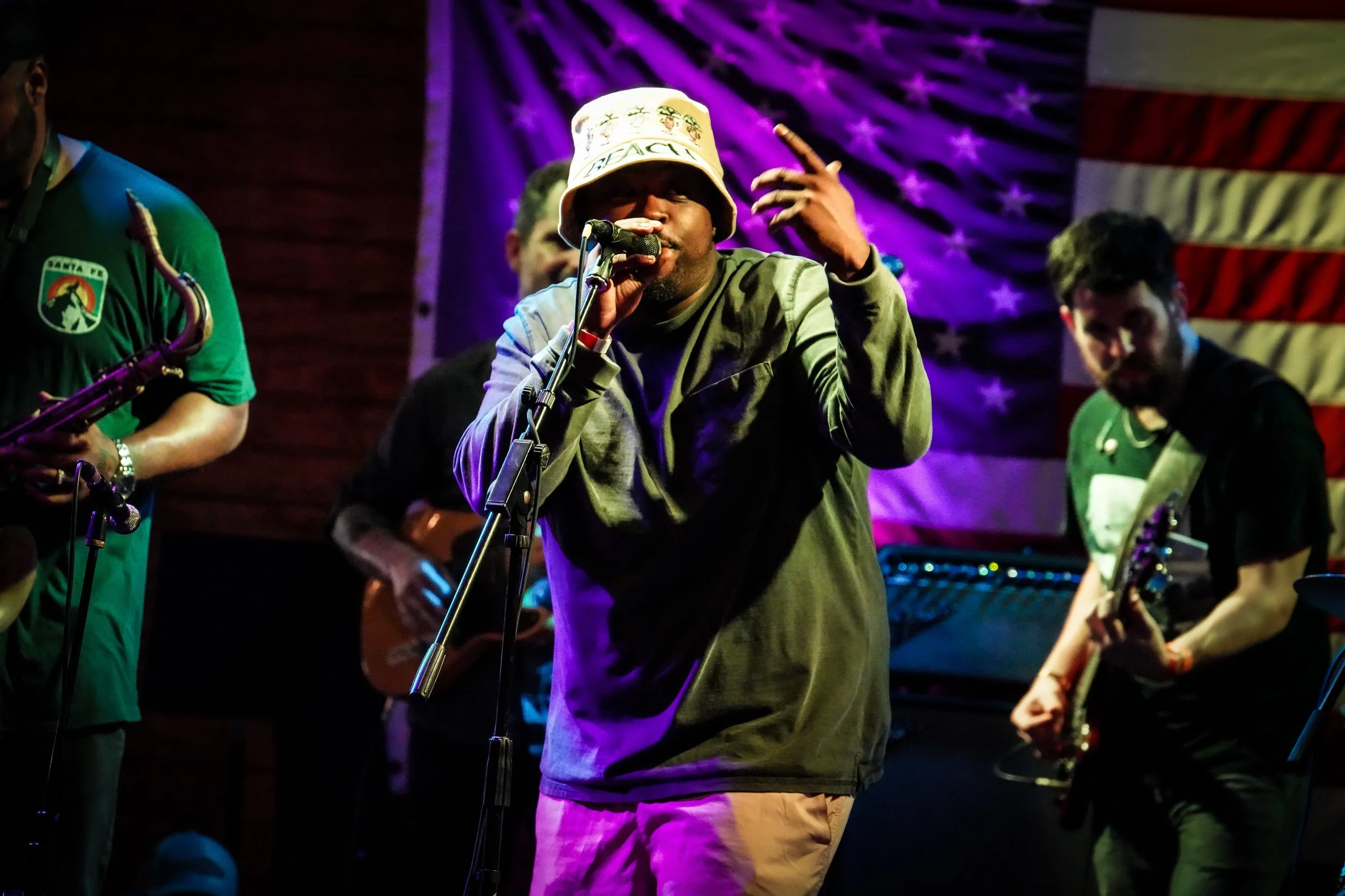 A musical performance with a singer in the center, wearing a bucket hat and gray jacket, singing into a microphone. Musicians around him play guitars and a saxophone, with a backdrop of an American flag and colorful lighting.