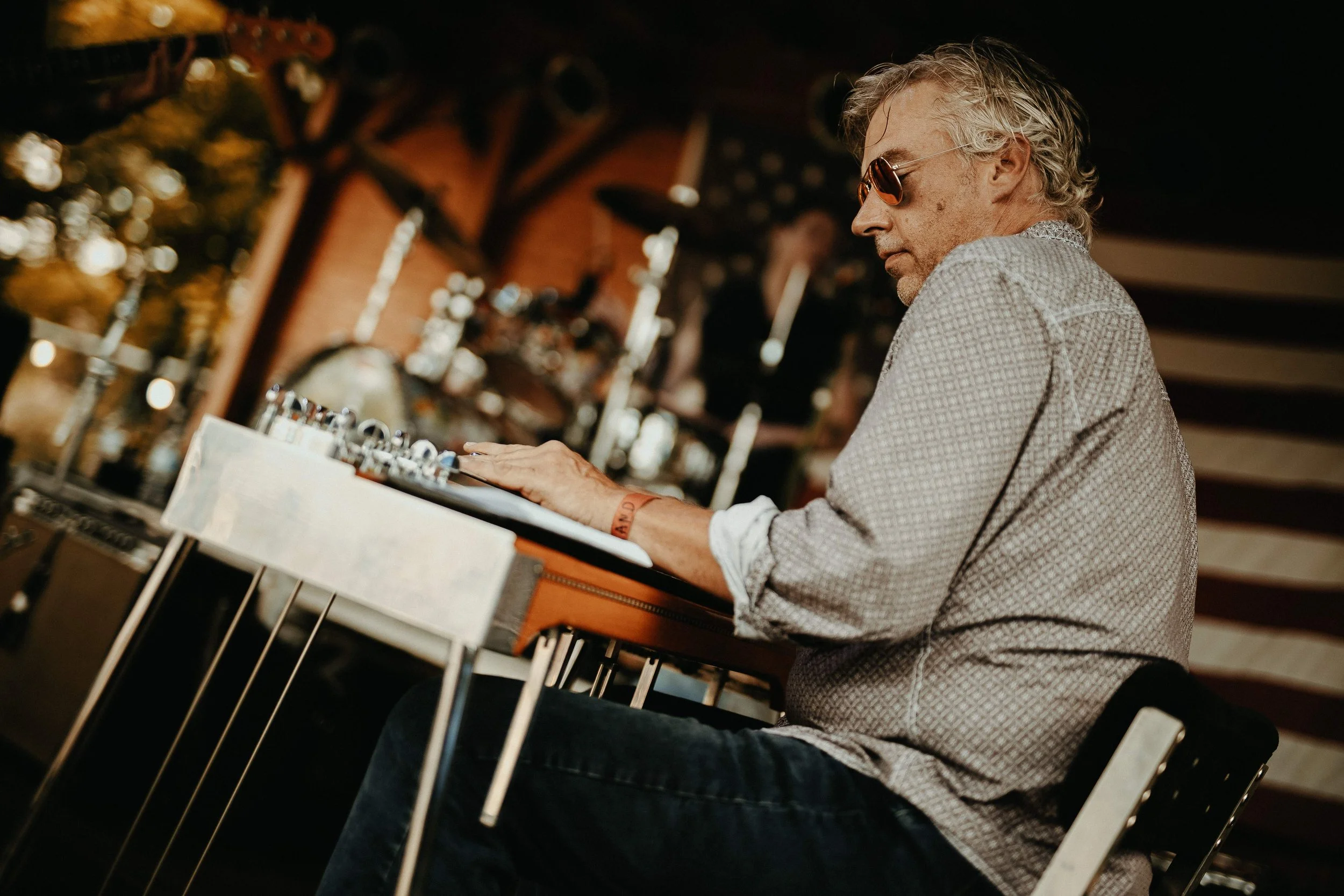 A man wearing sunglasses and a patterned shirt playing a vibraphone in a cozy, warmly lit room with musical instruments and decorations in the background.