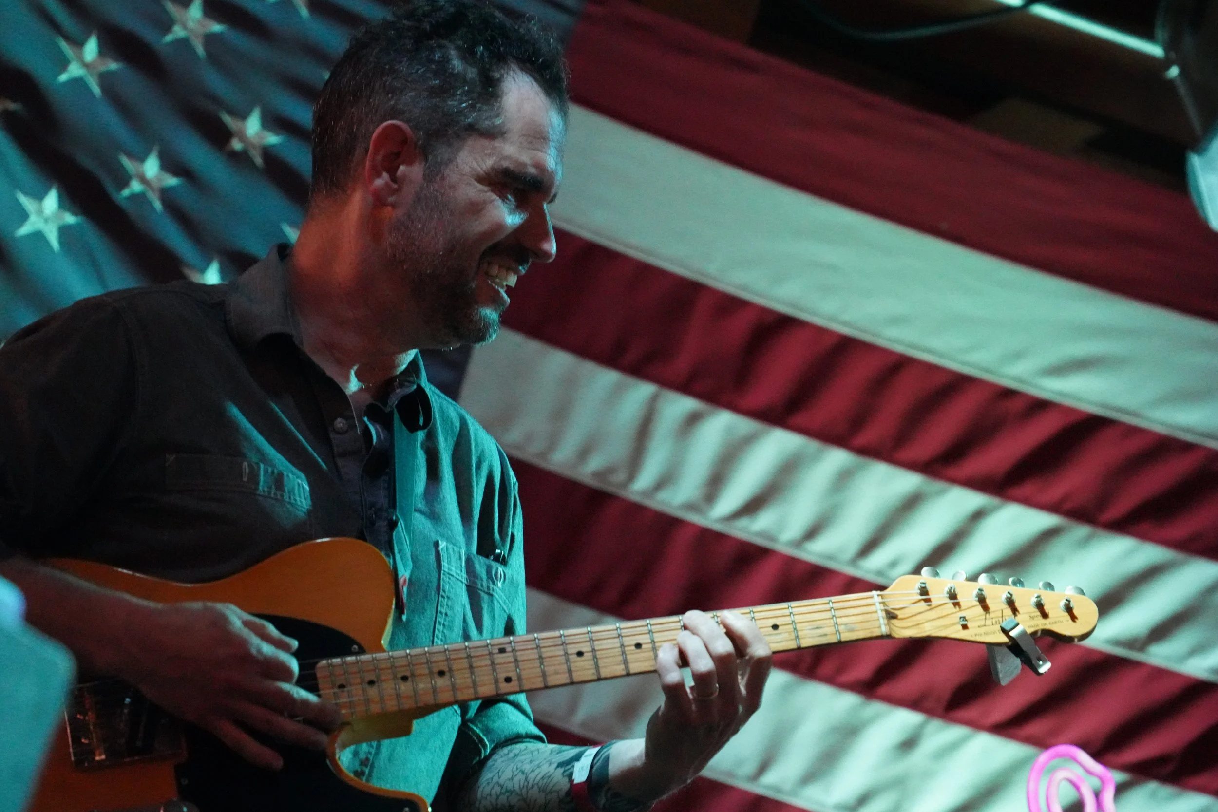 A man playing an electric guitar, smiling, with a patriotic American flag in the background.