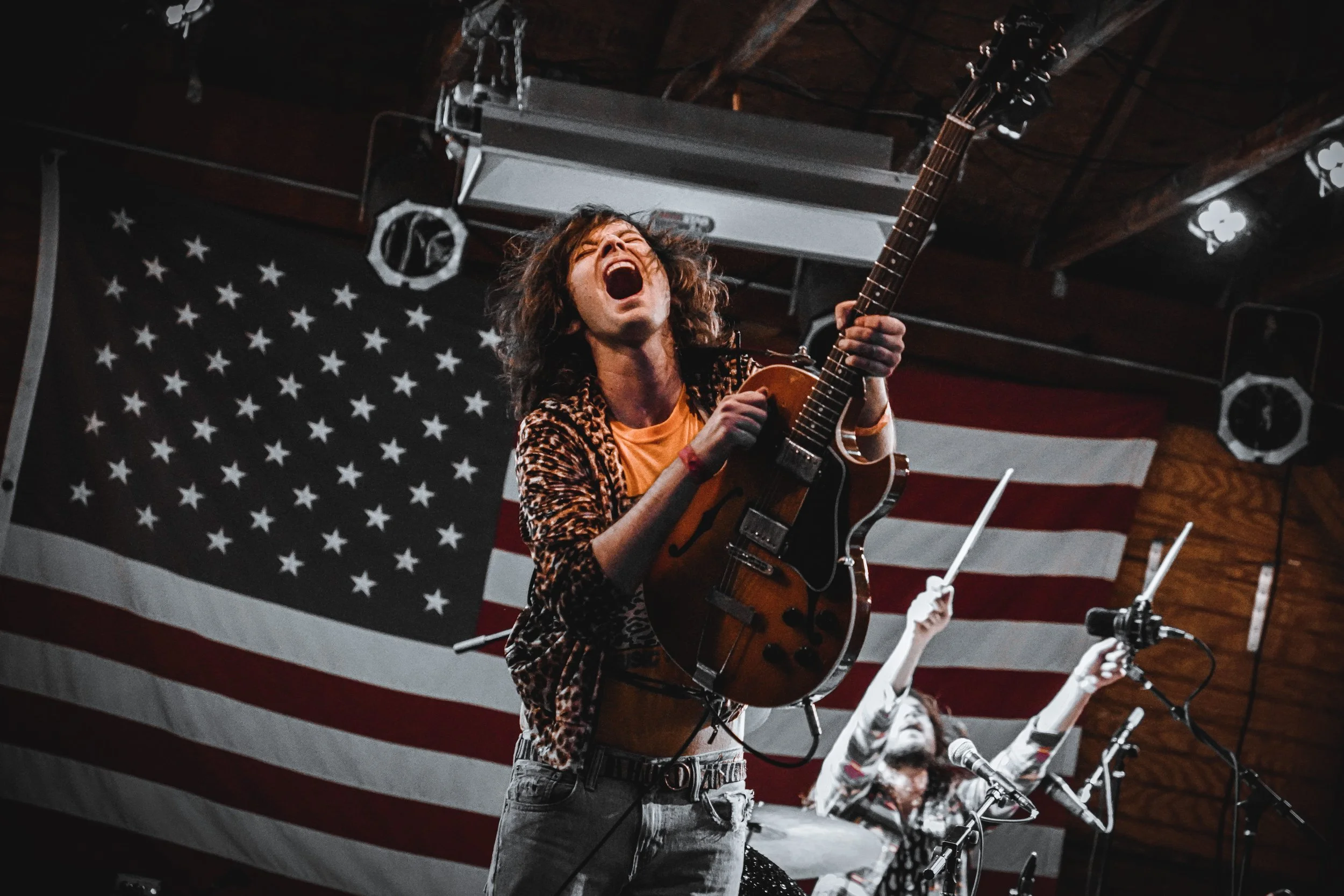 A musician with curly hair, wearing a leopard print shirt, passionately playing an electric guitar on stage with an American flag in the background.