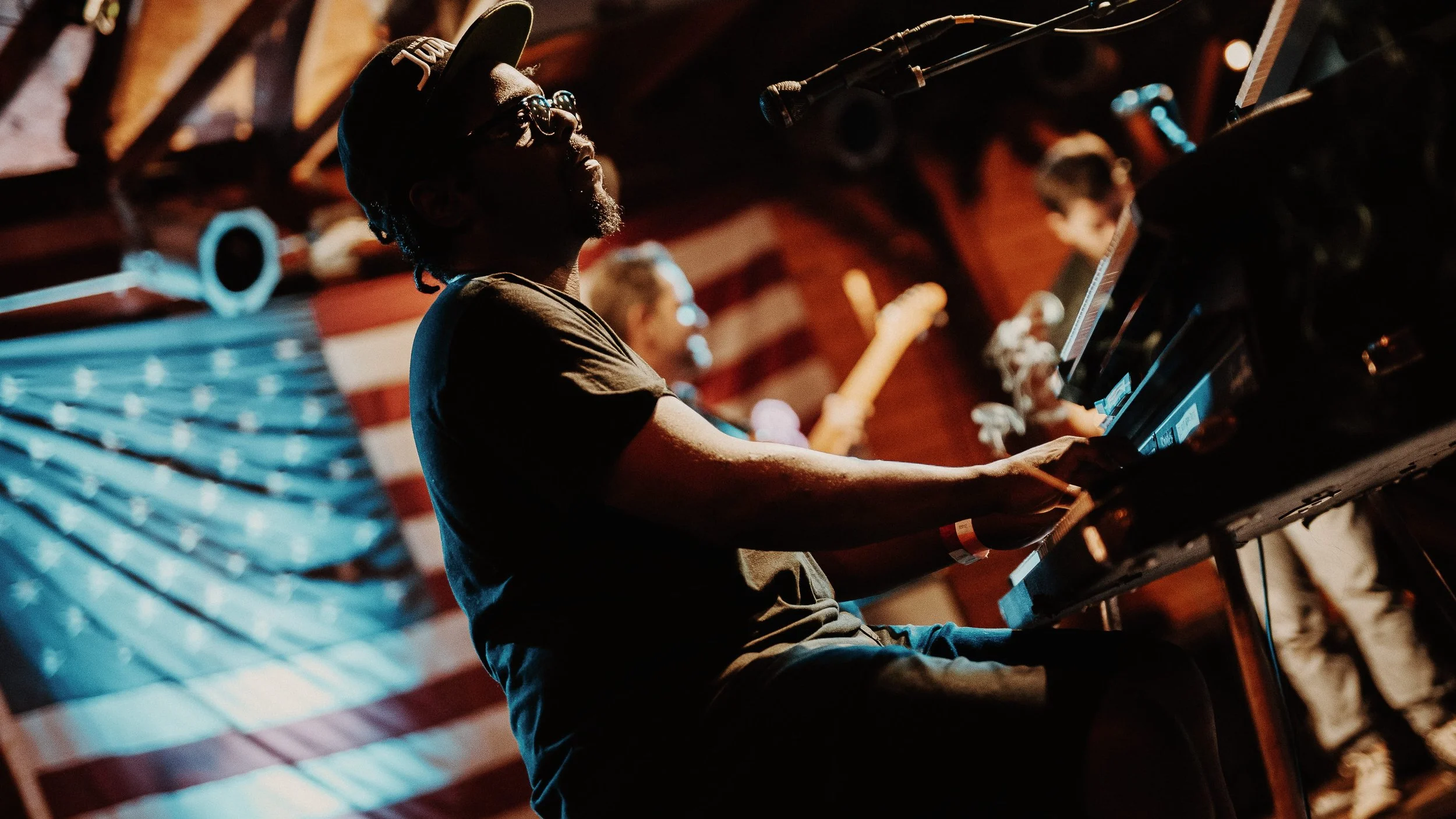 A musician playing the keyboard on stage, wearing a cap and glasses, with an American flag in the background.