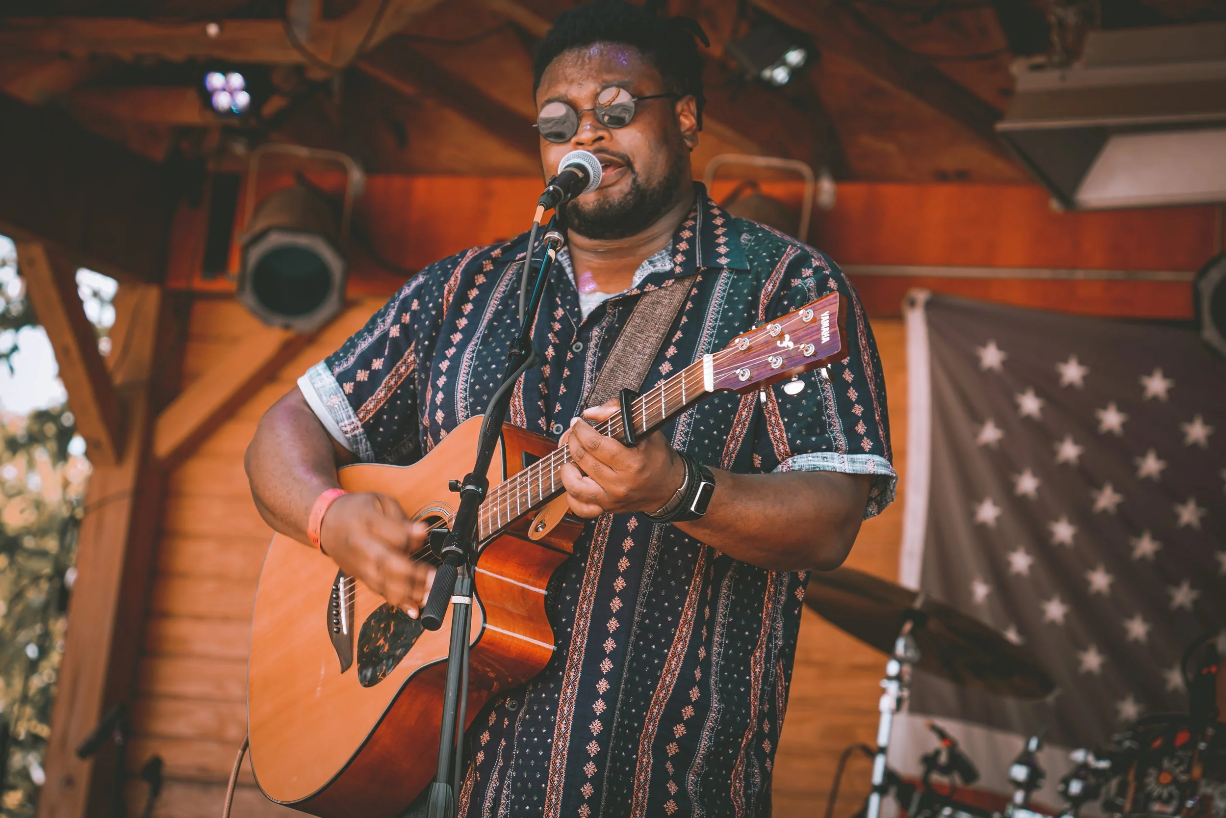 A man playing an acoustic guitar and singing into a microphone on a wooden stage with a patriotic banner in the background.