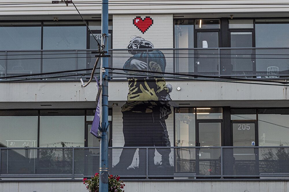 Mural of two people hugging with a cartoon style heart sitting above their heads - painted on a residential building between two balconies 