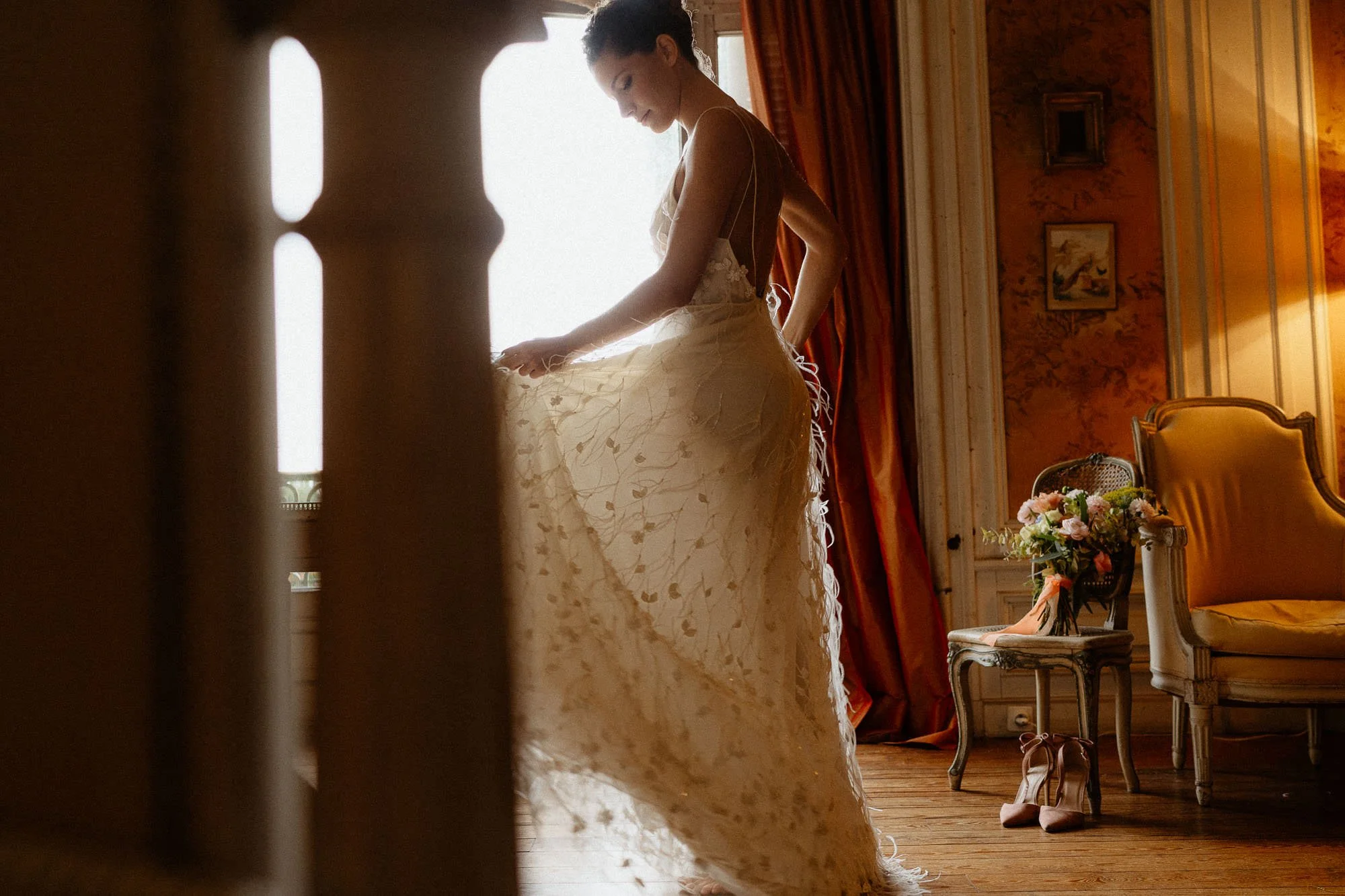 Une femme habillée en robe de mariée, regardant vers le sol, dans une pièce élégante avec des meubles anciens, une chaise avec un bouquet de fleurs, des chaussures à côté.