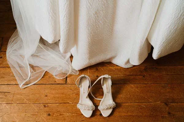 Des chaussures à talons blanches sous une table recouverte d'une draperie blanche sur un sol en bois.