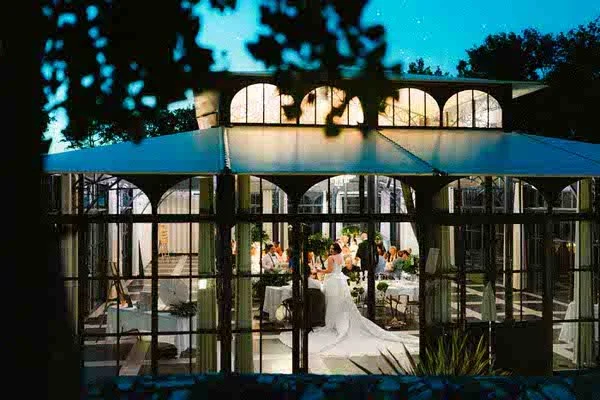 Vue d'une réception ou d'un mariage à l'intérieur d'une verrière ou d'une serre, avec des invités assis autour de tables, sous un grand parasol, vue à travers une porte ou une fenêtre.