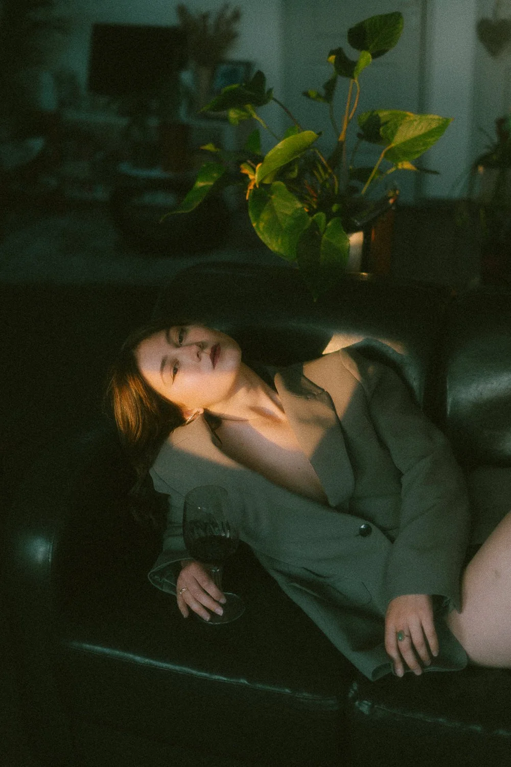 Une femme assise dans un fauteuil, tenant un verre de vin rouge, dans un environnement intérieur sombre avec une grande plante verte à l'arrière.