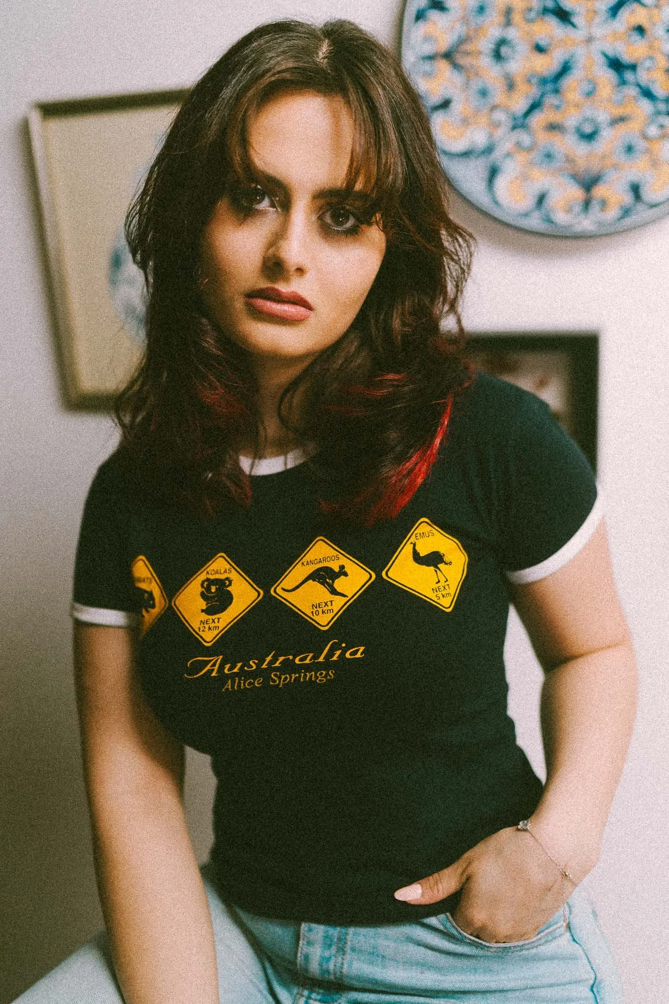 Une jeune femme aux cheveux bruns avec des reflets rouges, portant un t-shirt noir avec des illustrations de panneaux routiers australiens, assise sur une surface blanche, avec un tableau et un objet décoratif en arrière-plan.