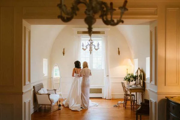 Deux femmes en robe de mariée se tenant par la taille dans une pièce élégante avec un chandelier au plafond, un miroir, un fauteuil, une table avec des fleurs et un lampadaire, avec une grande fenêtre laissant entrer la lumière.