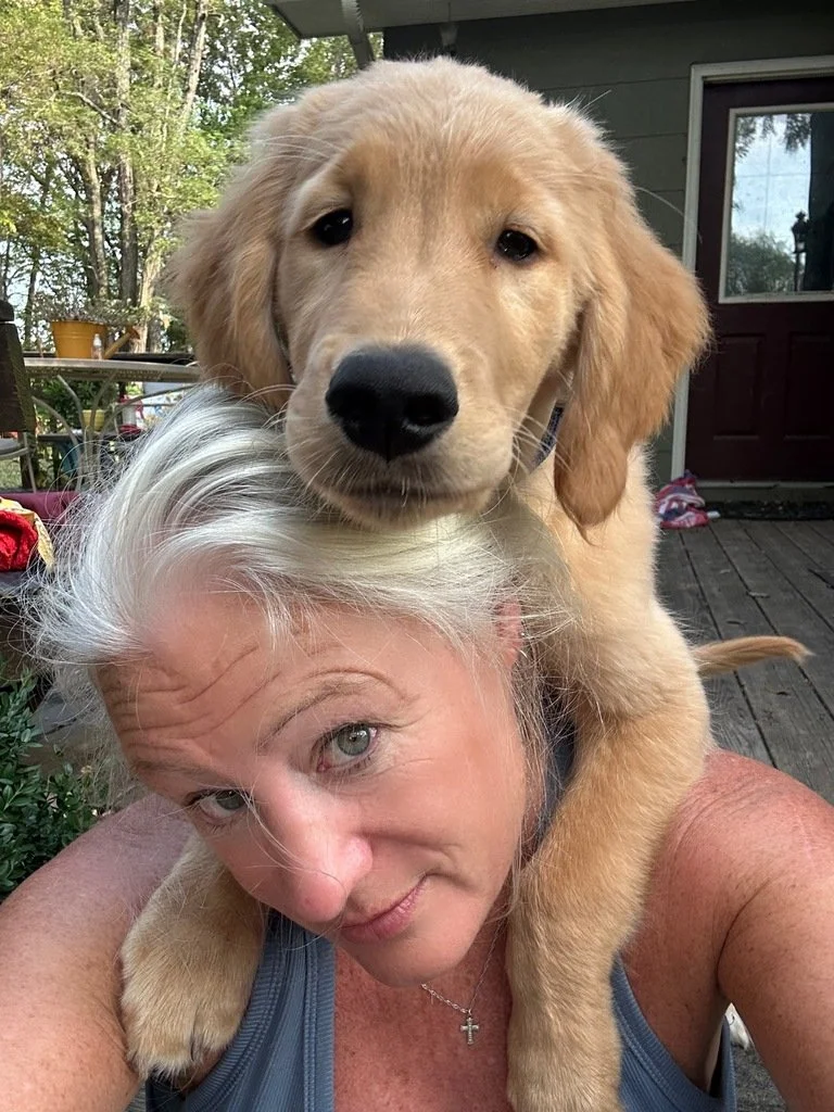 A woman with gray hair and green eyes taking a selfie with a golden retriever puppy resting on her shoulders outdoors.