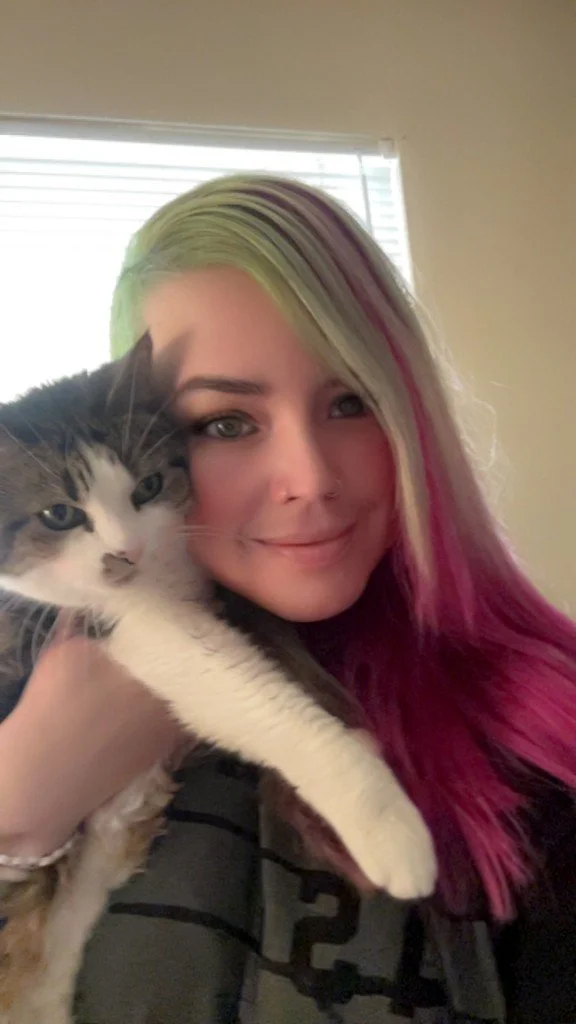 A young woman with colorful hair (green, purple, and pink) smiling and holding a fluffy gray and white cat close to her face, indoors near a window.